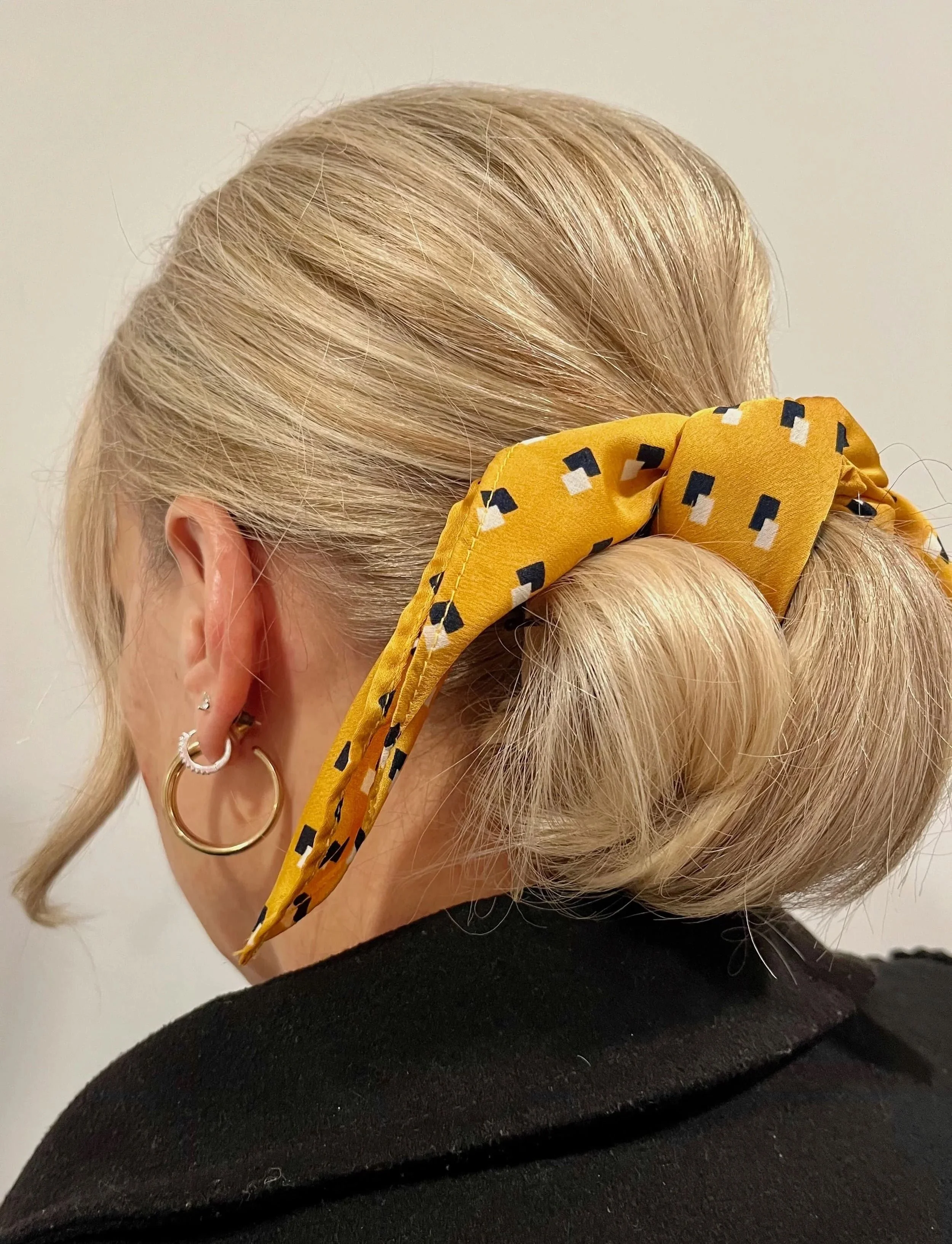 A woman with blonde hair styled in a low bun, wearing earrings, and a yellow headscarf with black and white patterns tied around her hair.