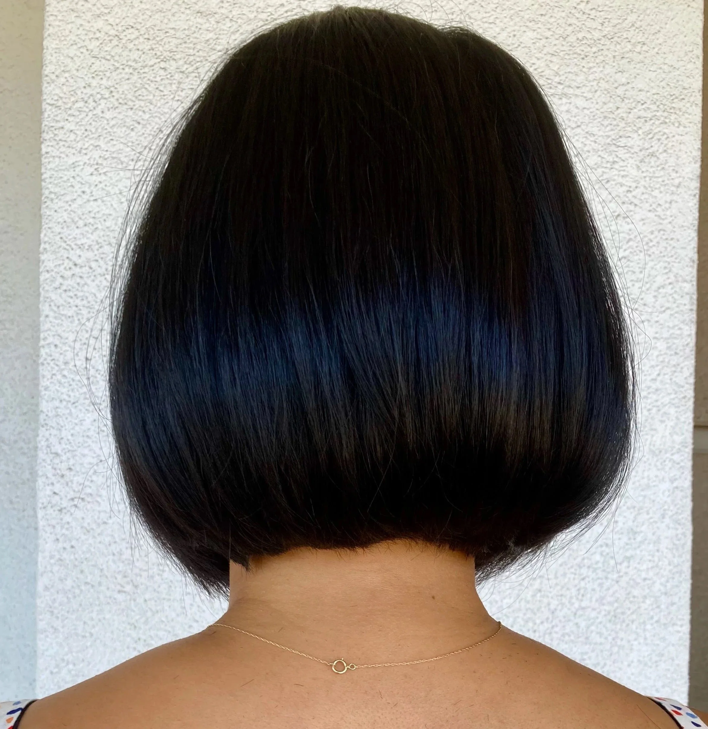 Back view of a woman with a short, sleek black bob haircut, wearing a thin gold necklace, and standing against a textured light-colored wall.
