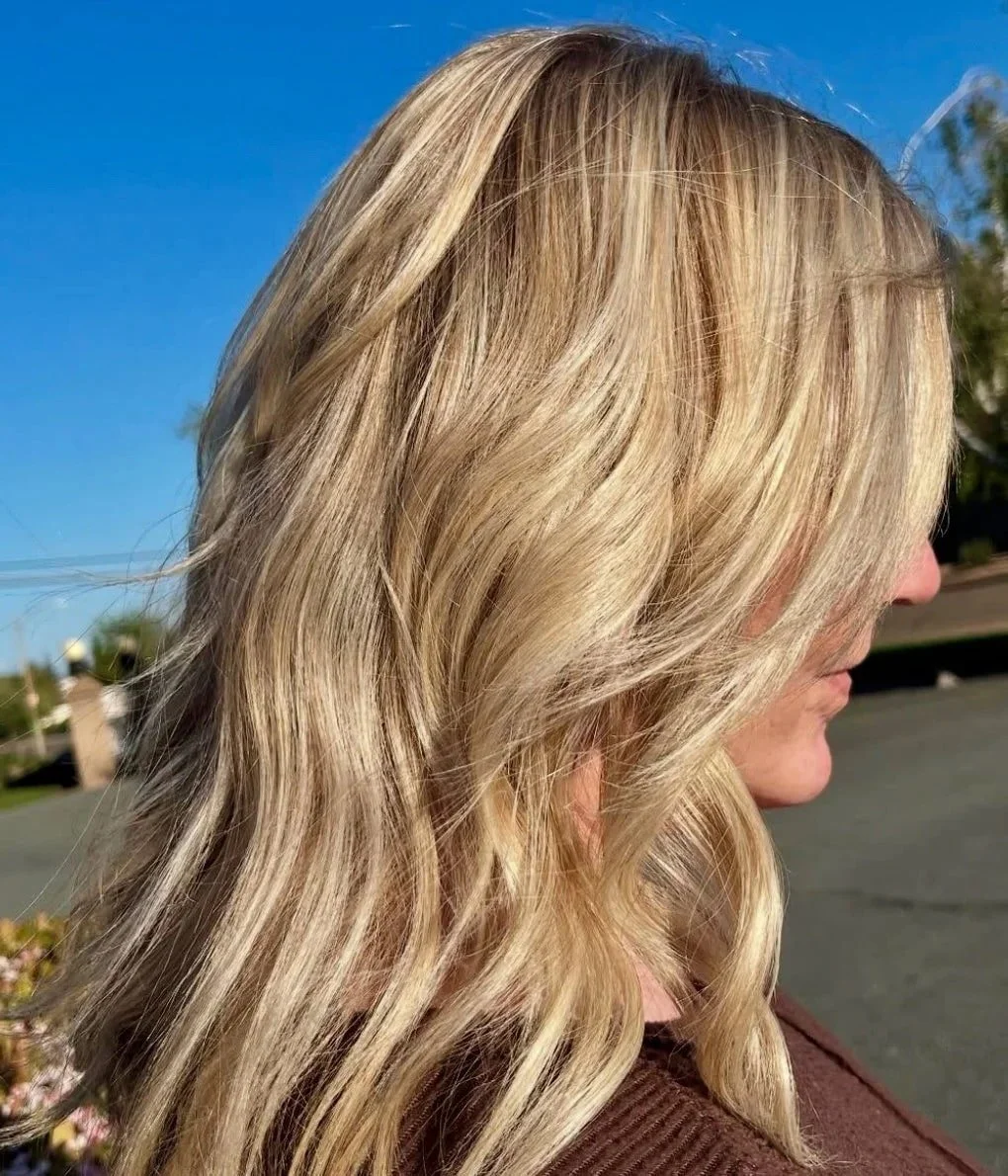 Close-up of a woman with long, wavy blonde hair standing outdoors on a sunny day with a clear blue sky in the background.