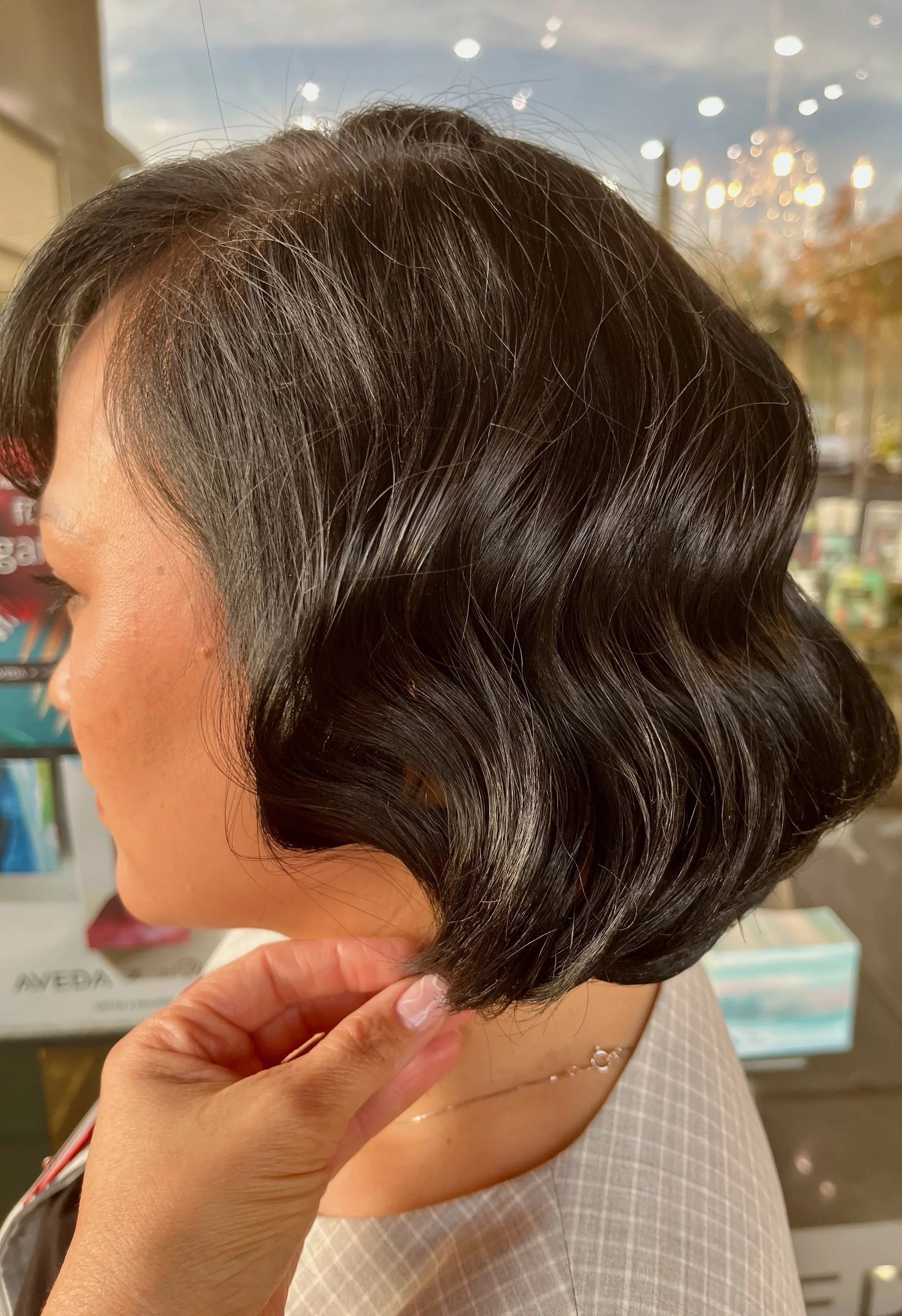 Close-up of a woman with a freshly finger wave styled short, wavy black haircut, looking downward while someone gently holds her hair. The background shows a store window with outdoor reflections and store products.