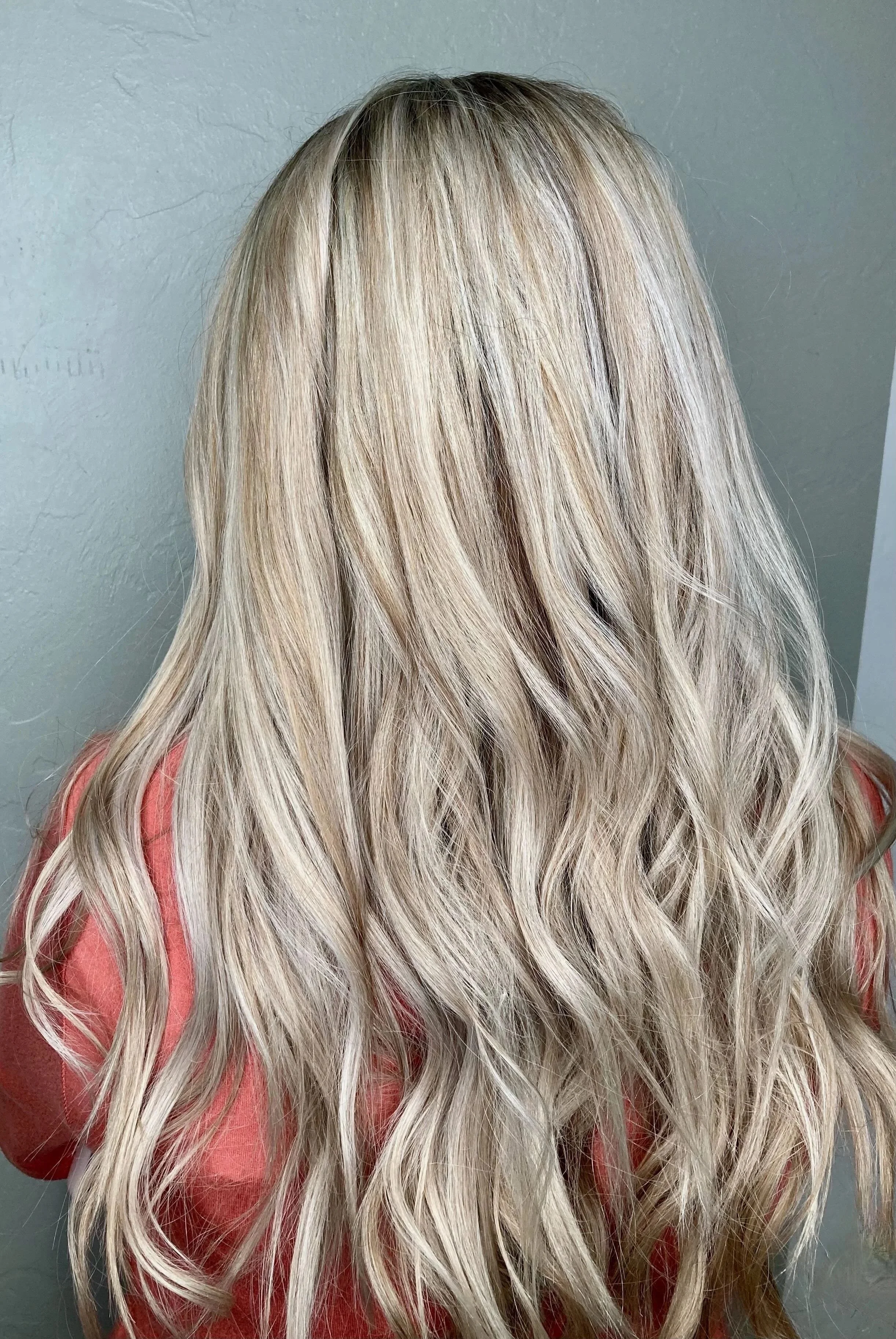 Back view of a person with long, wavy blonde hair, wearing a coral-colored top, against a plain gray wall.
