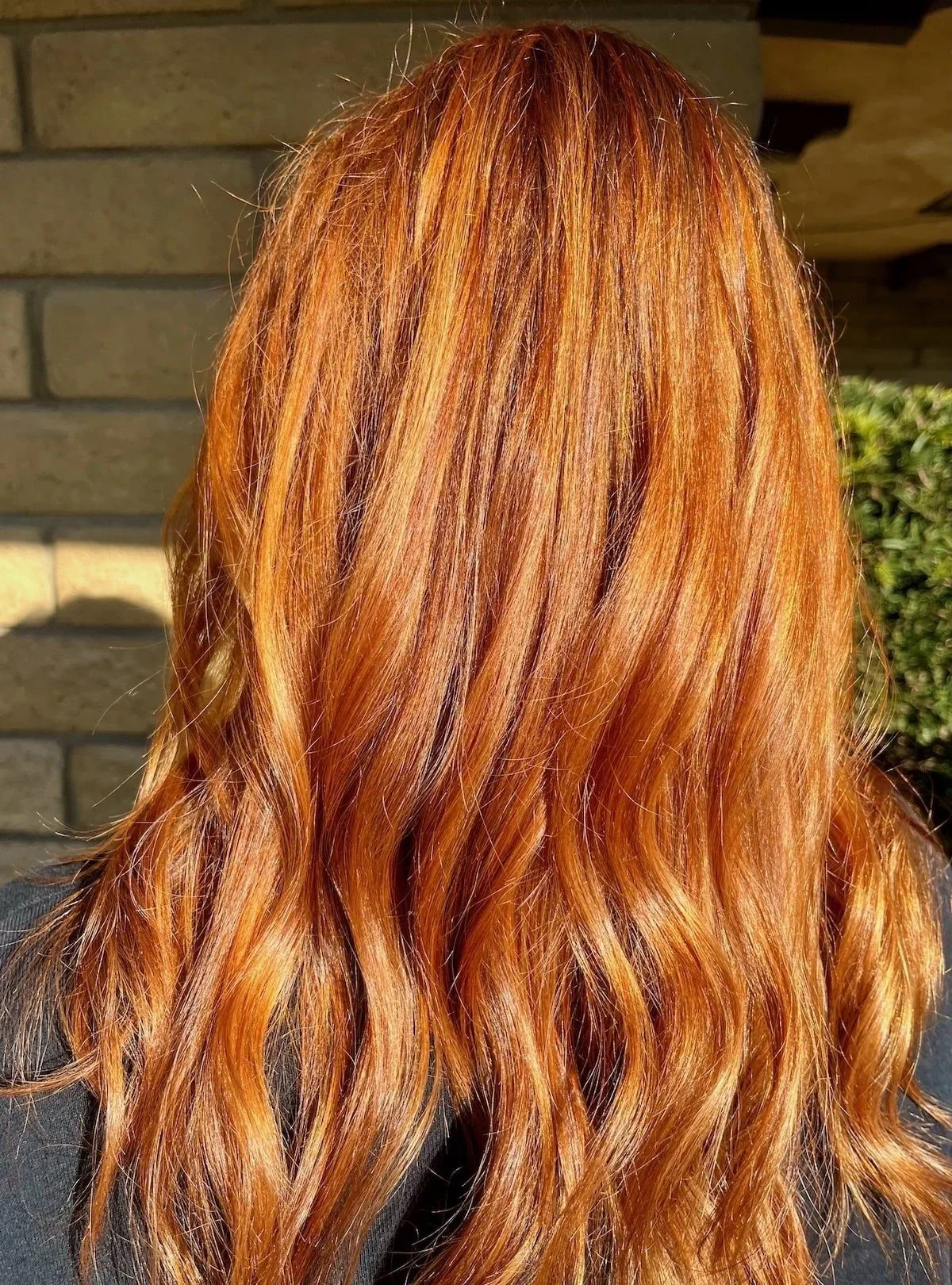 Back view of a person with long, wavy, red hair standing outdoors near a brick wall and some greenery.
