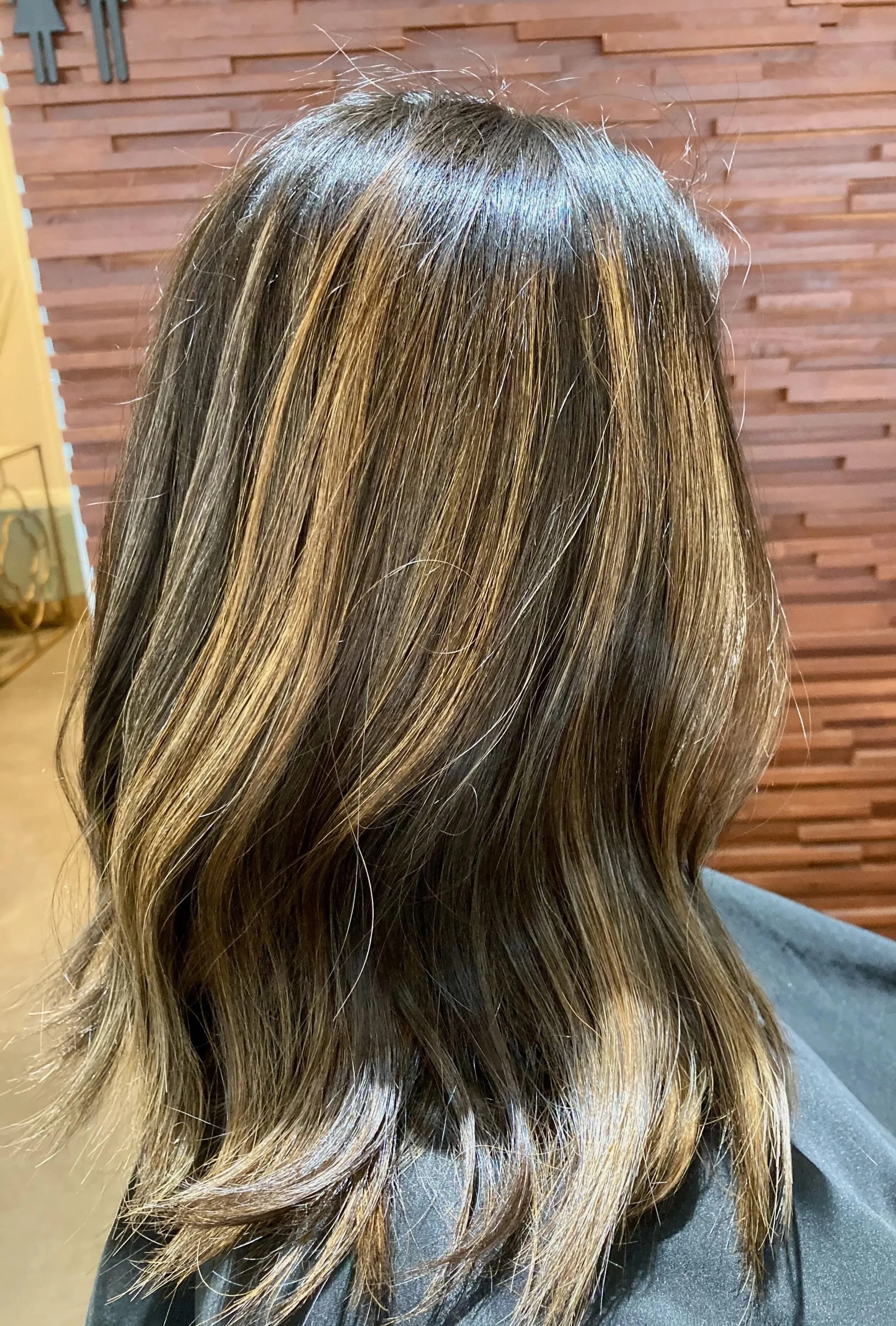 A woman's medium-length, wavy hair with light brown and blonde highlights, viewed from the back against a wooden wall background.