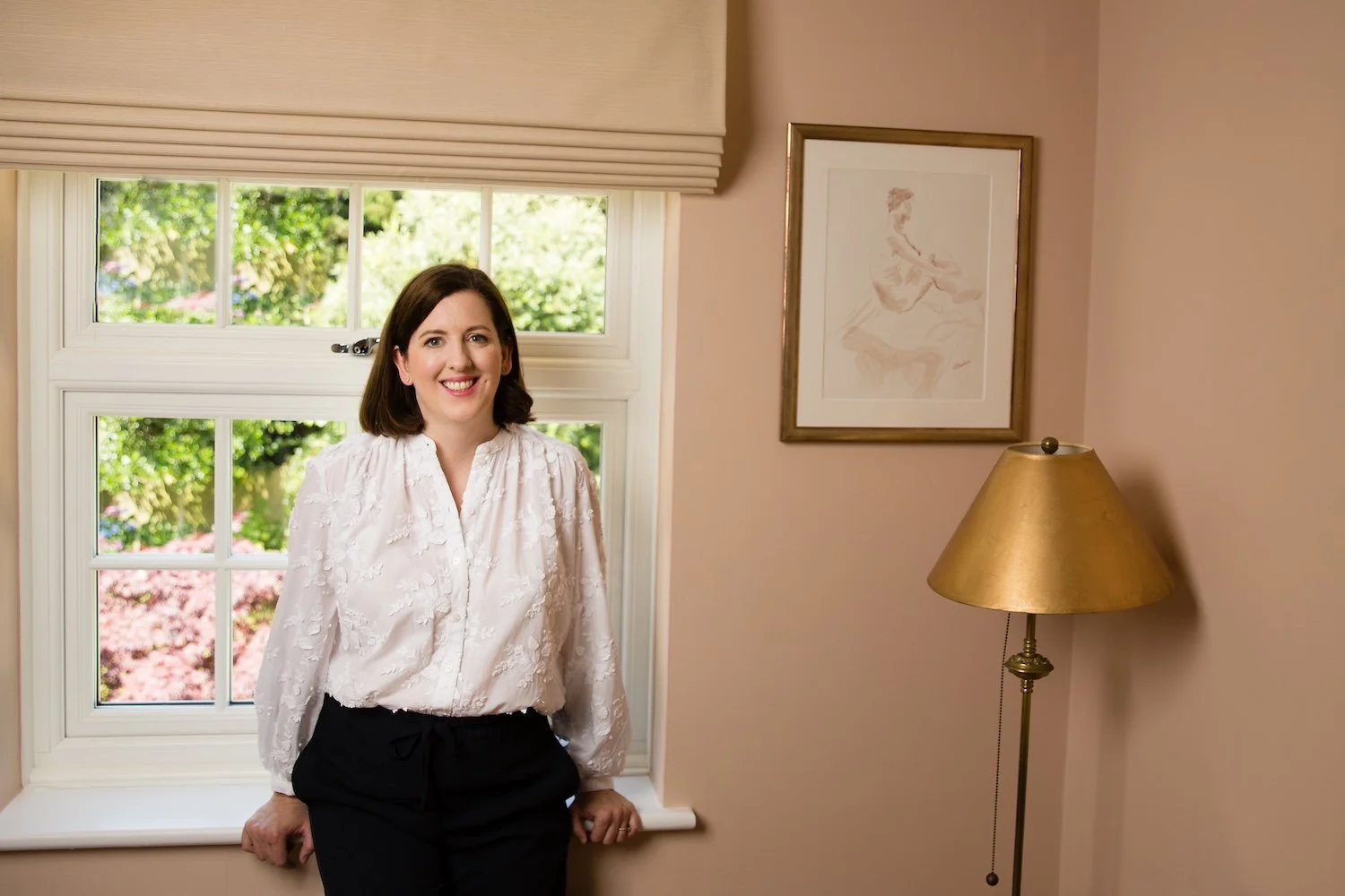 Laura is smiling, standing in front of a window with a view of trees and pink flowers, in a well decorated room with beige walls, a framed artwork, and a gold floor lamp.