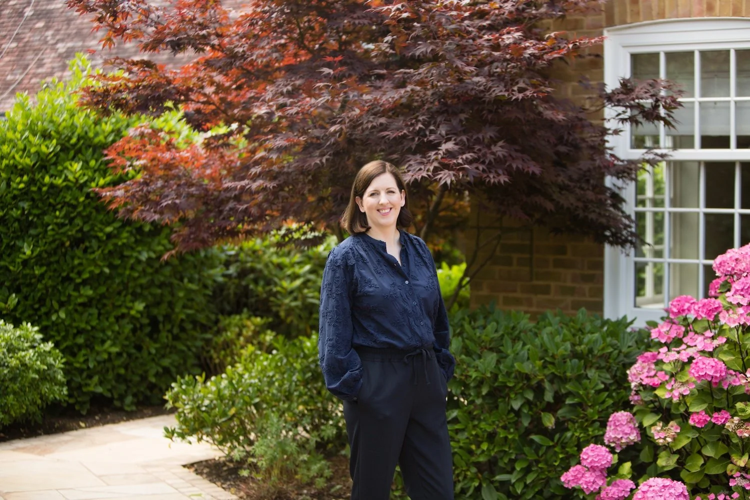 Laura standing outdoors in a garden with pink hydrangeas, green bushes, and a red-leafed tree, next to a house with a large window based near Guildford, Surrey.
