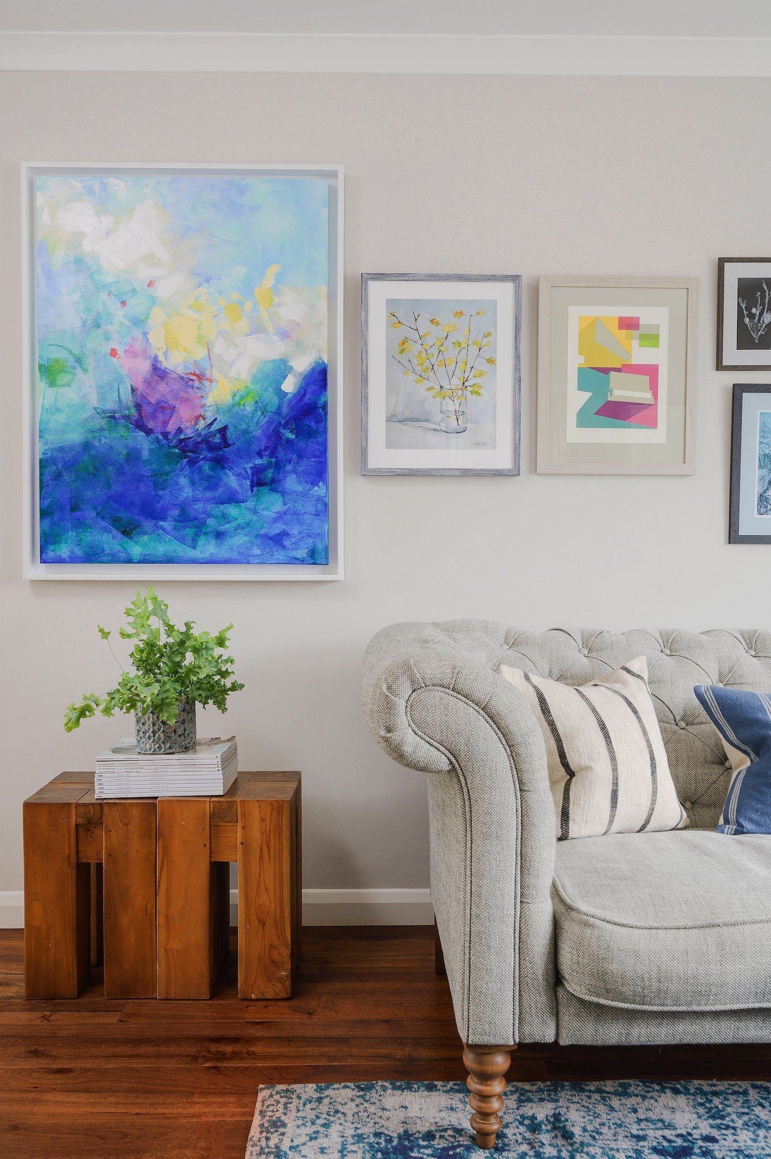 A living room with optimal interior design for someone with colour blindness. There is a beige sofa with striped pillows, a wooden side table with a green plant and books, and a wall decorated with framed artwork in various sizes and styles.