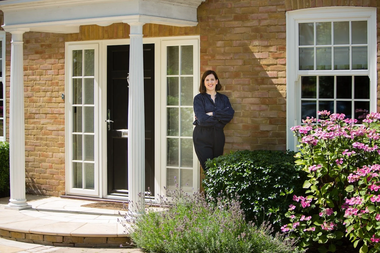 Laura is wearing a navy blouse and black trousers, standing in front of a Surrey house she has completed an interior design project on. She is on a porch with white columns in front of a brick house with white-framed windows and a black door.