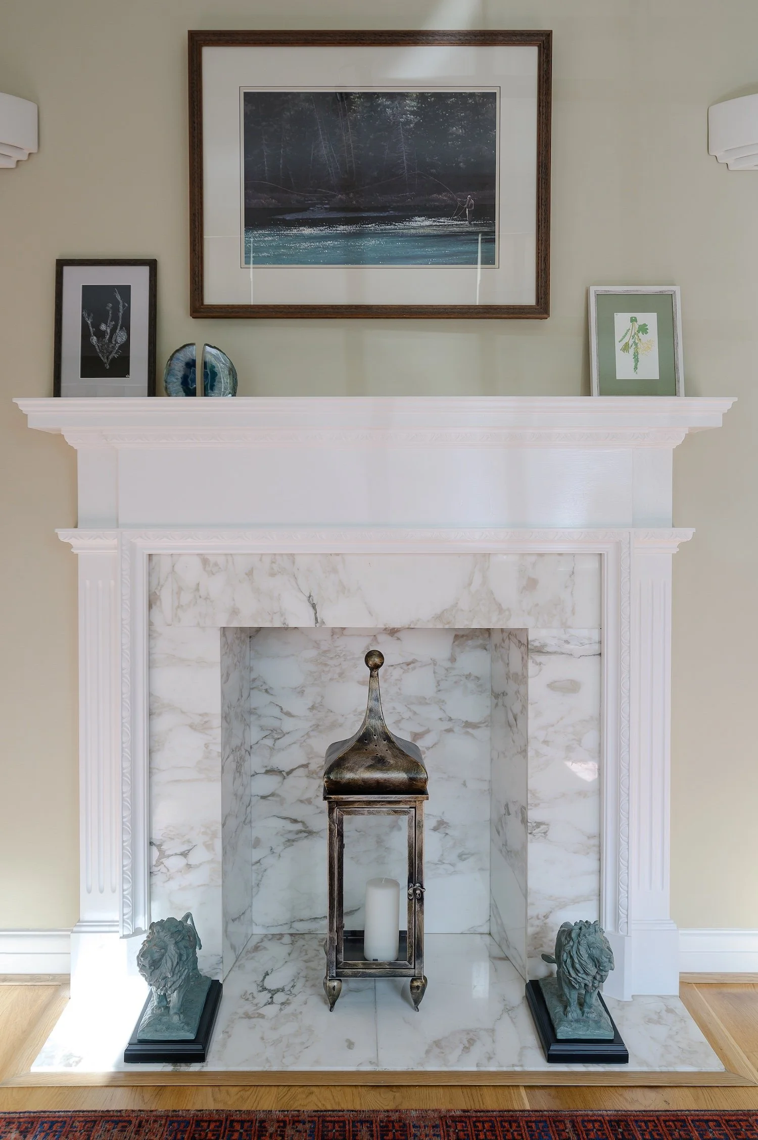 A front room designed by Laura, in Surrey. Decorative white marble fireplace with a metal lantern and white candle inside, flanked by two lion head sculptures on black bases. Framed artwork and decorative objects are on the mantle, and wall.