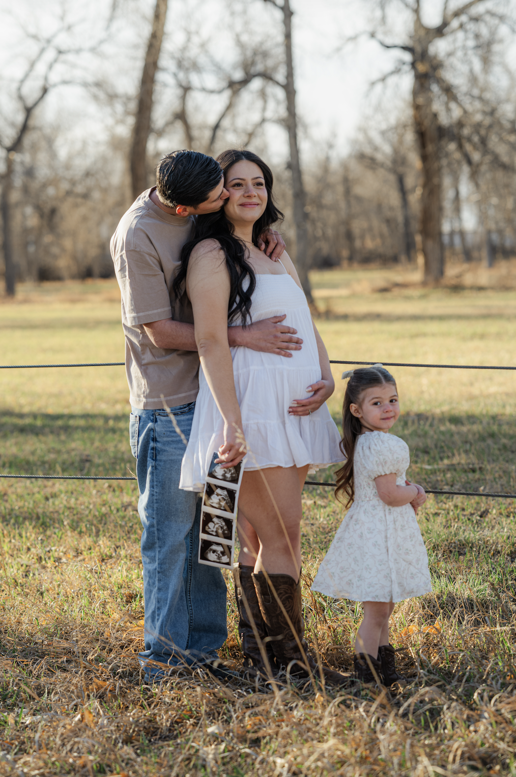 A pregnant woman standing outdoors with her partner and young daughter, holding ultrasound photos, during golden sunset in a grassy field with trees. Shot by PhotosByMaya Kirschenbaum