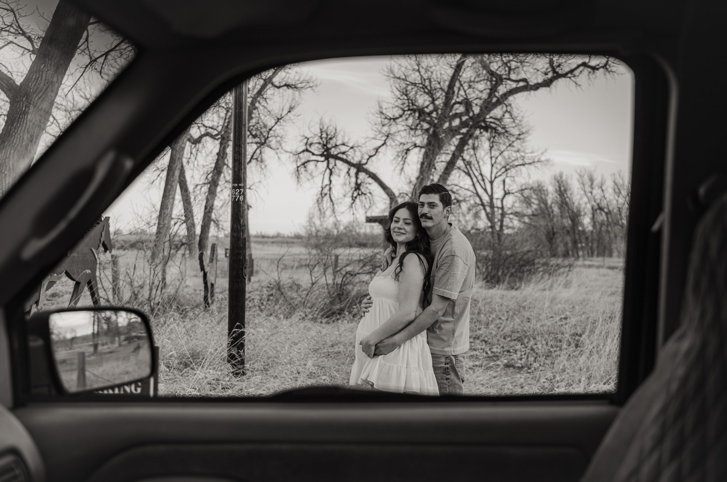 A man and woman viewed from the inside of a vehicle, with trees and grass in the background, with a black and white maternity shoot in Colorado. 