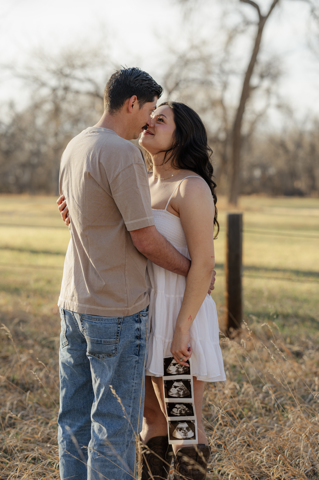 A couple standing close together outdoors in Denver, Colorado embracing each other with a scenic background landscape of trees and grass, holding ultrasound images, indicating they are expecting a baby.