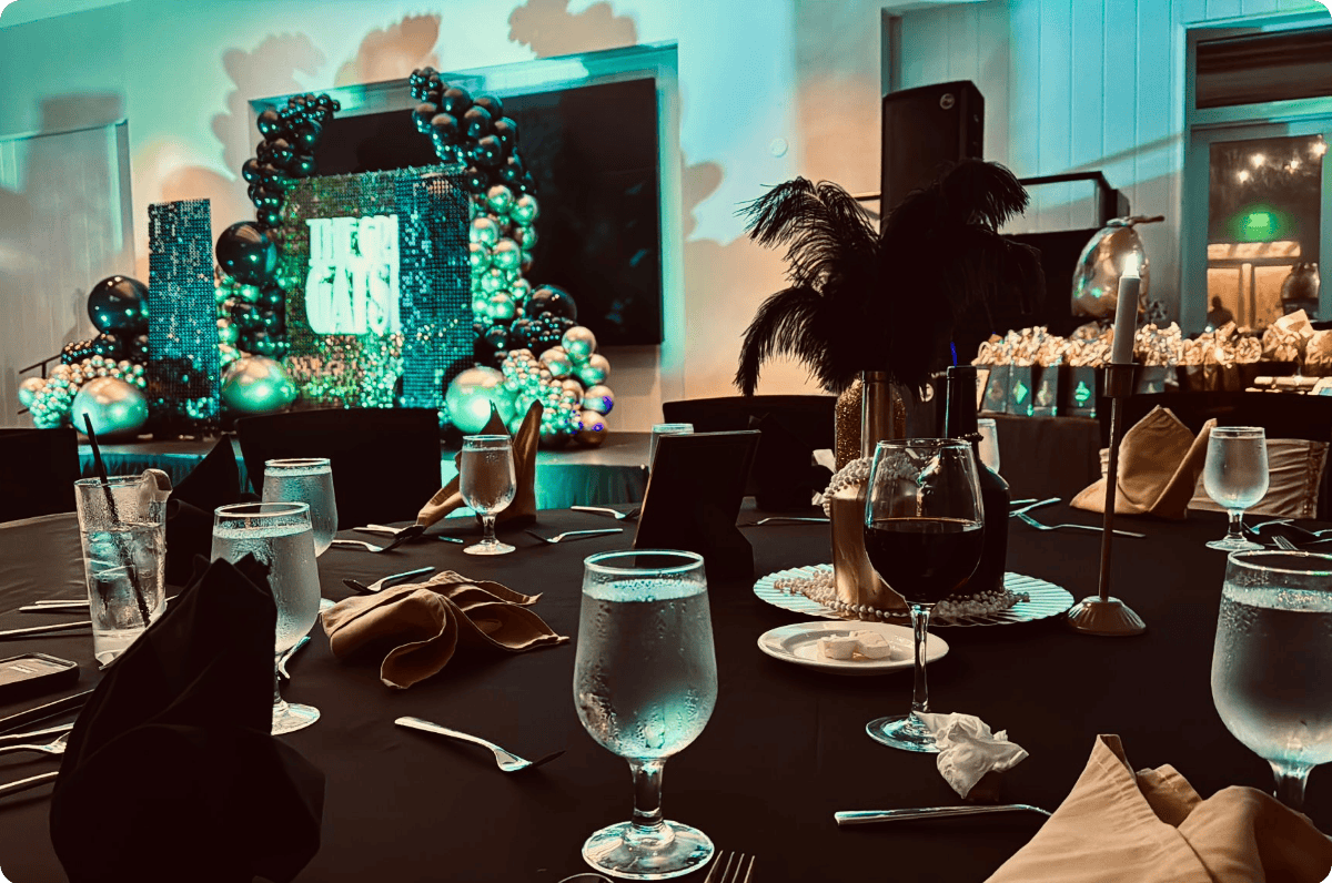 Decorated event table with glasses of water, a wine glass, napkins, and a lit candle, with a large black feather centerpiece and an ornate backdrop with balloons and sign reading 'The Gas' in the background.