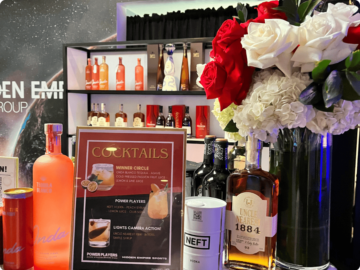 A bar setup with various bottles of alcohol, a large bouquet of red and white flowers, and a cocktail menu. The menu features drink options such as Winner Circle, Power Players, and Lights Camera Action.