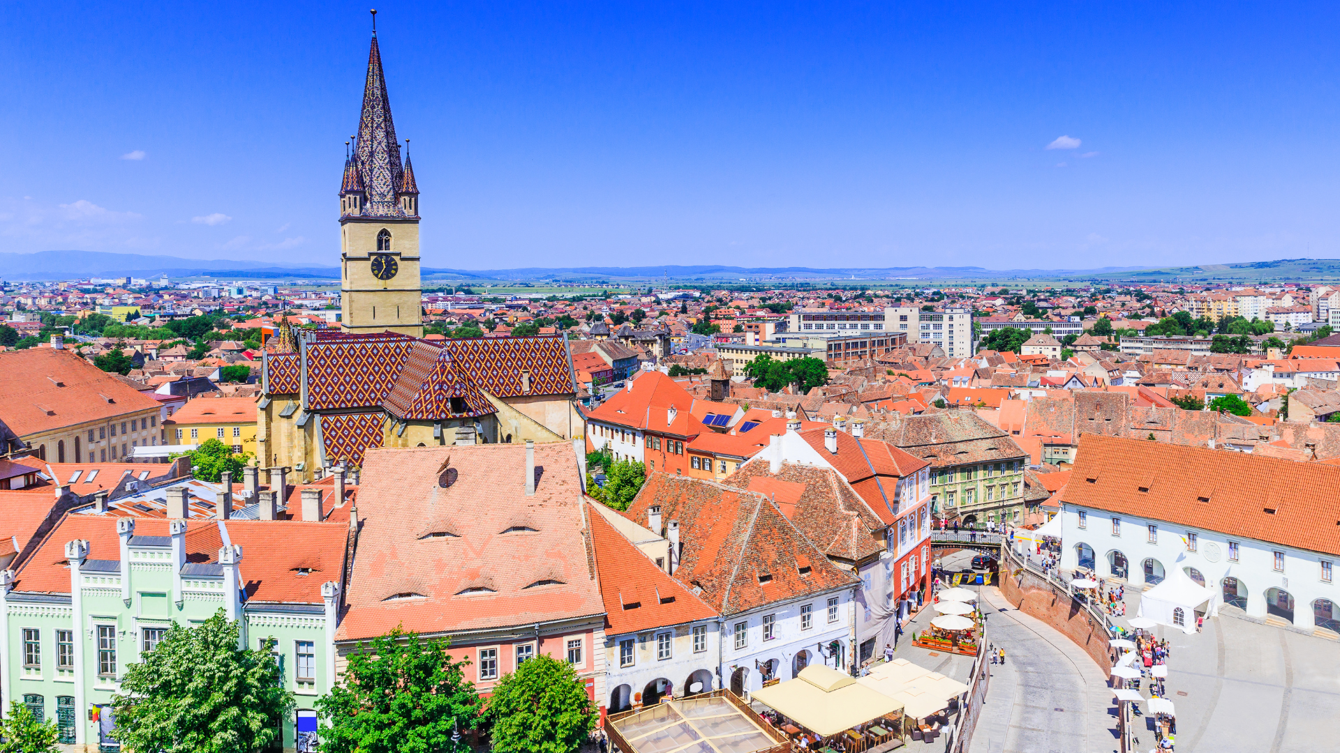 A panoramic view of a European city with historic buildings, a church with a tall spire featuring colorful patterned roof tiles, around a busy town square with outdoor cafes and market stalls, under a bright blue sky with a few clouds.