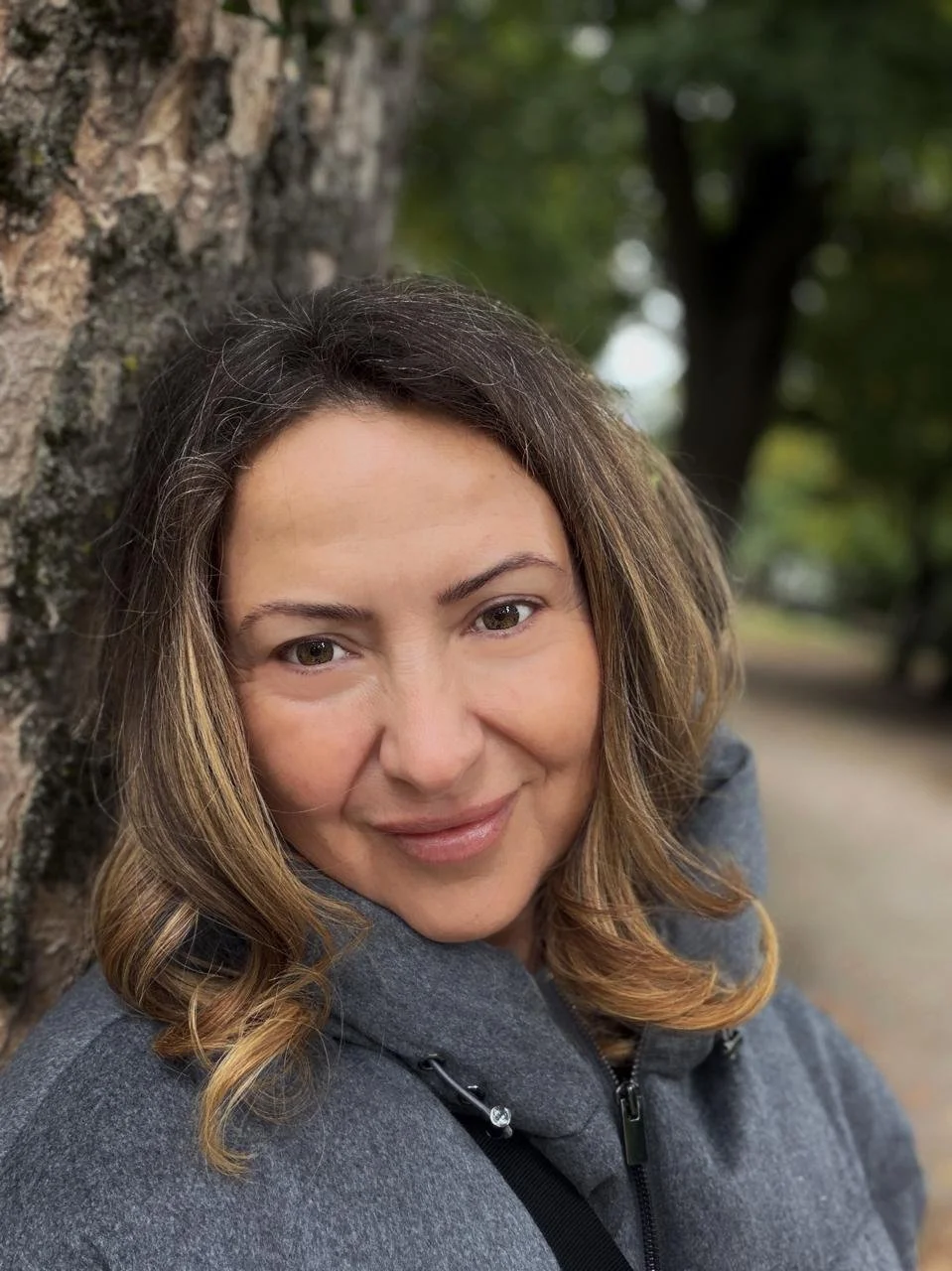 A woman with wavy brown hair and light skin leaning against a tree in a park.
