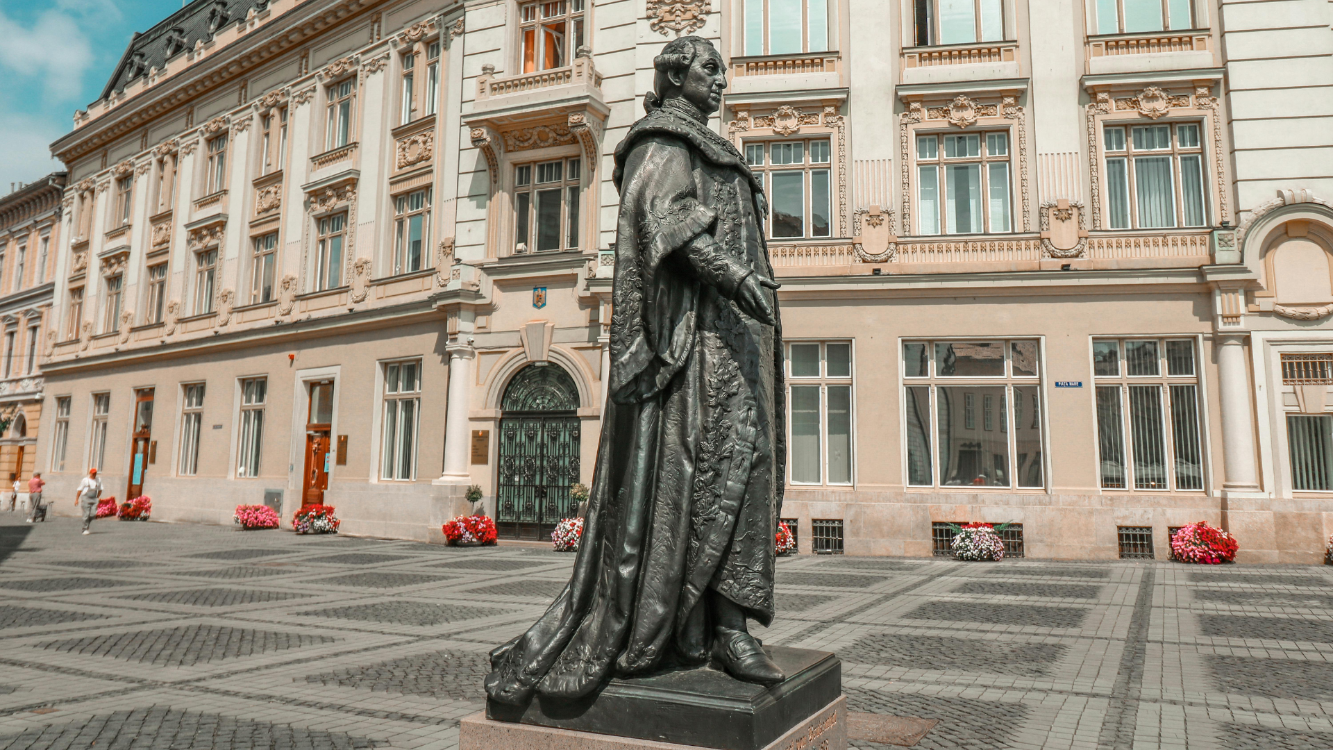 Bronze statue of a historical figure in medieval attire, standing outdoors in front of an ornate building with multiple windows and decorative architectural details, surrounded by flower arrangements.
