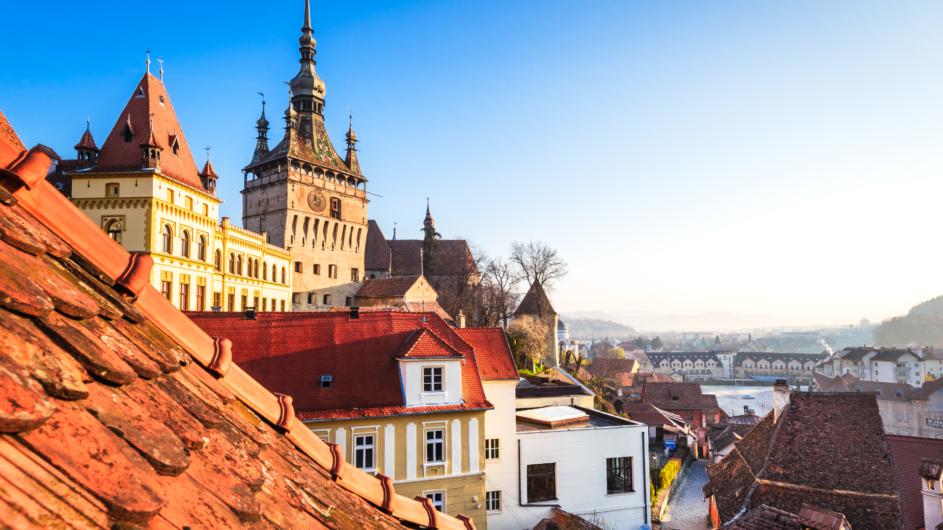 A scenic view of a historic European town with colorful buildings and a castle with towers, red-tiled roofs, and a river in the background under a clear blue sky.