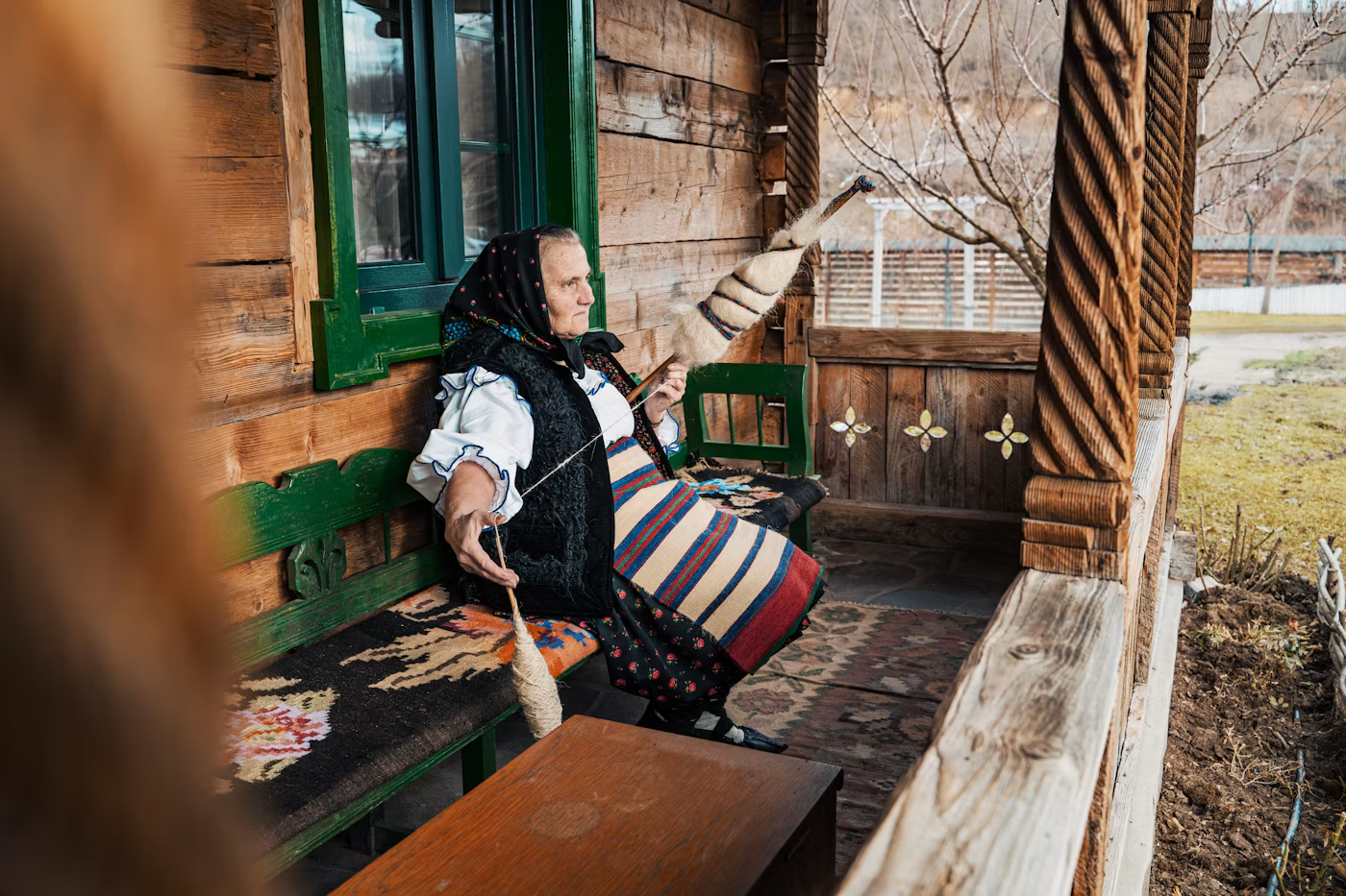 Elderly woman dressed in traditional folk clothing sitting on a wooden bench on a porch, knitting a sock on her lap, with a handmade sock on her raised arm, outside a rustic wooden house.