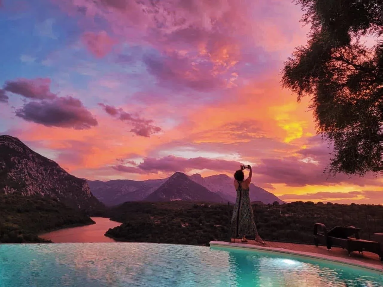 A woman in a patterned dress is taking a photo with her phone beside a pool, overlooking a landscape of mountains and a river at sunset with colorful clouds in the sky.