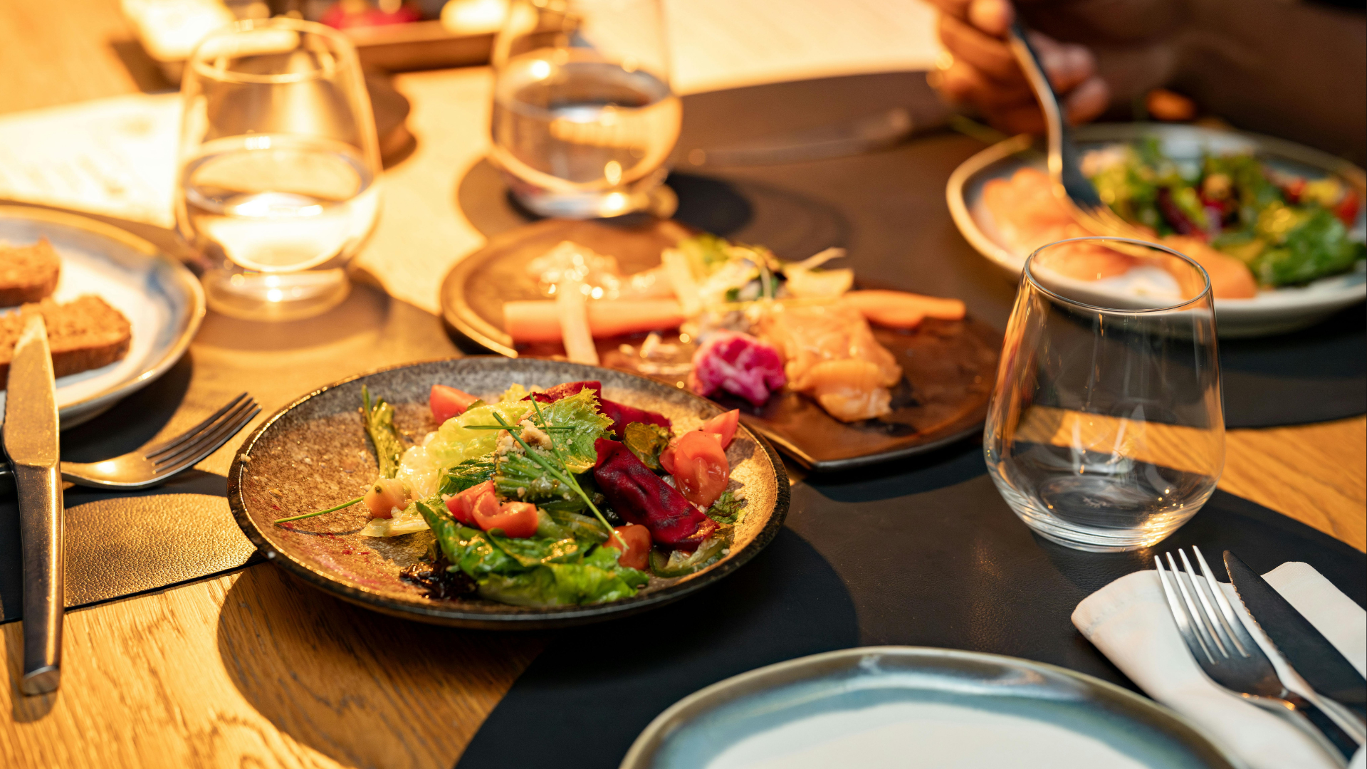 A table set for a meal with plates of salad, sliced meat, bread, and glasses of water, illuminated with warm lighting.