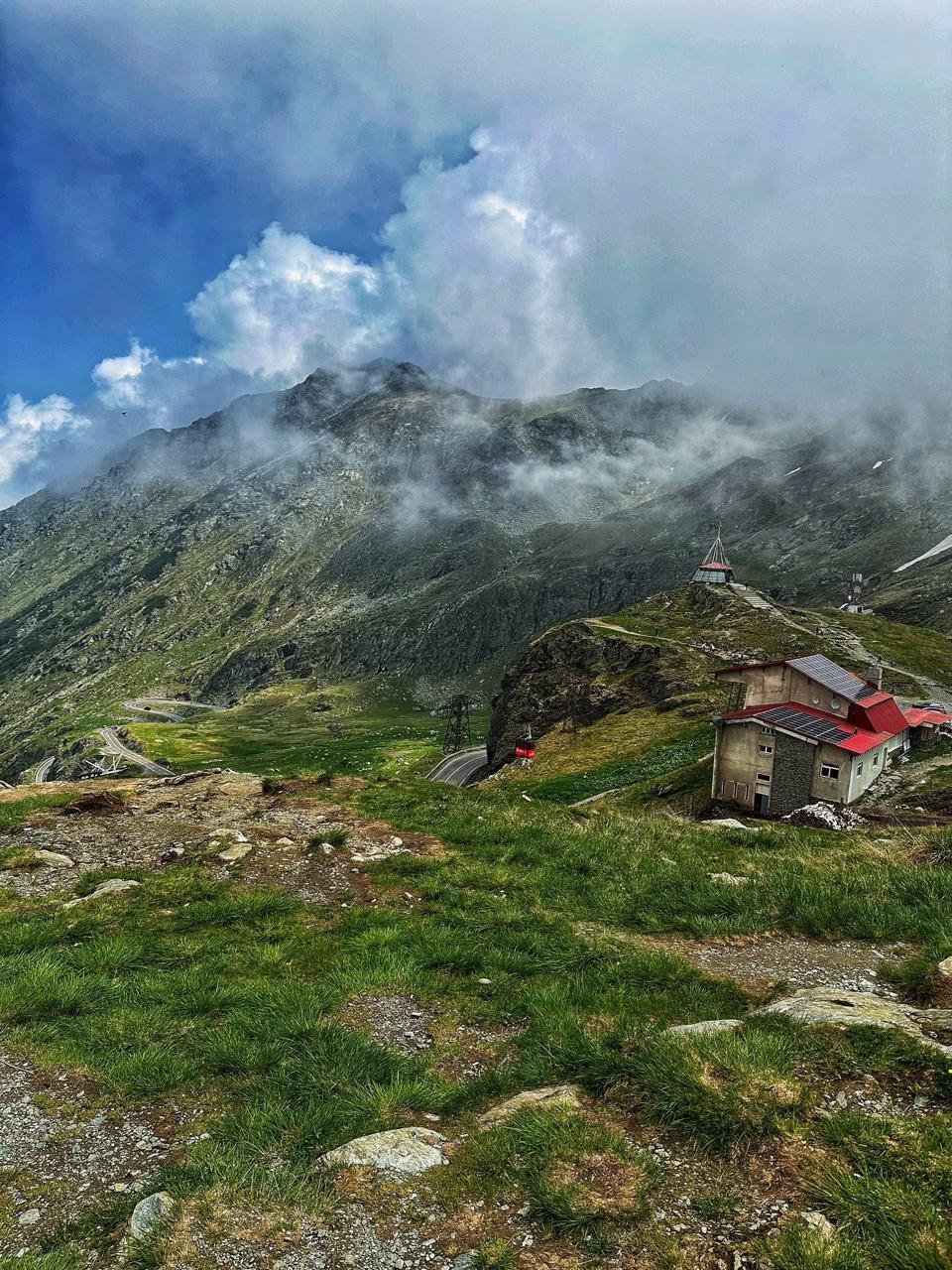 Mountain landscape with green grassy foreground, winding roads, and a few small buildings with red and gray roofs. Mist and clouds surround the mountain peaks in the background.