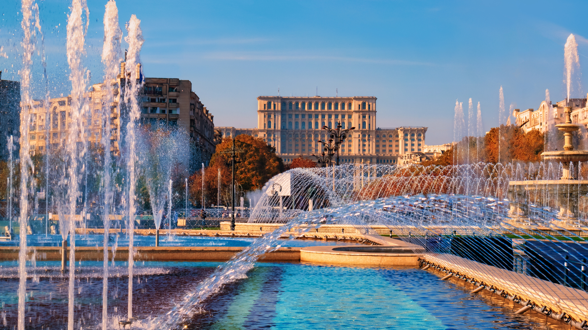 Fountain with water jets in front of a large historic building under a blue sky.