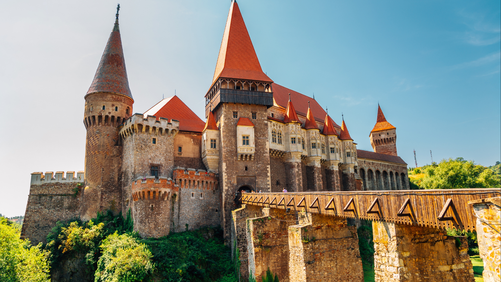 A medieval castle with tall towers and red-tiled roofs, surrounded by greenery against a clear blue sky.