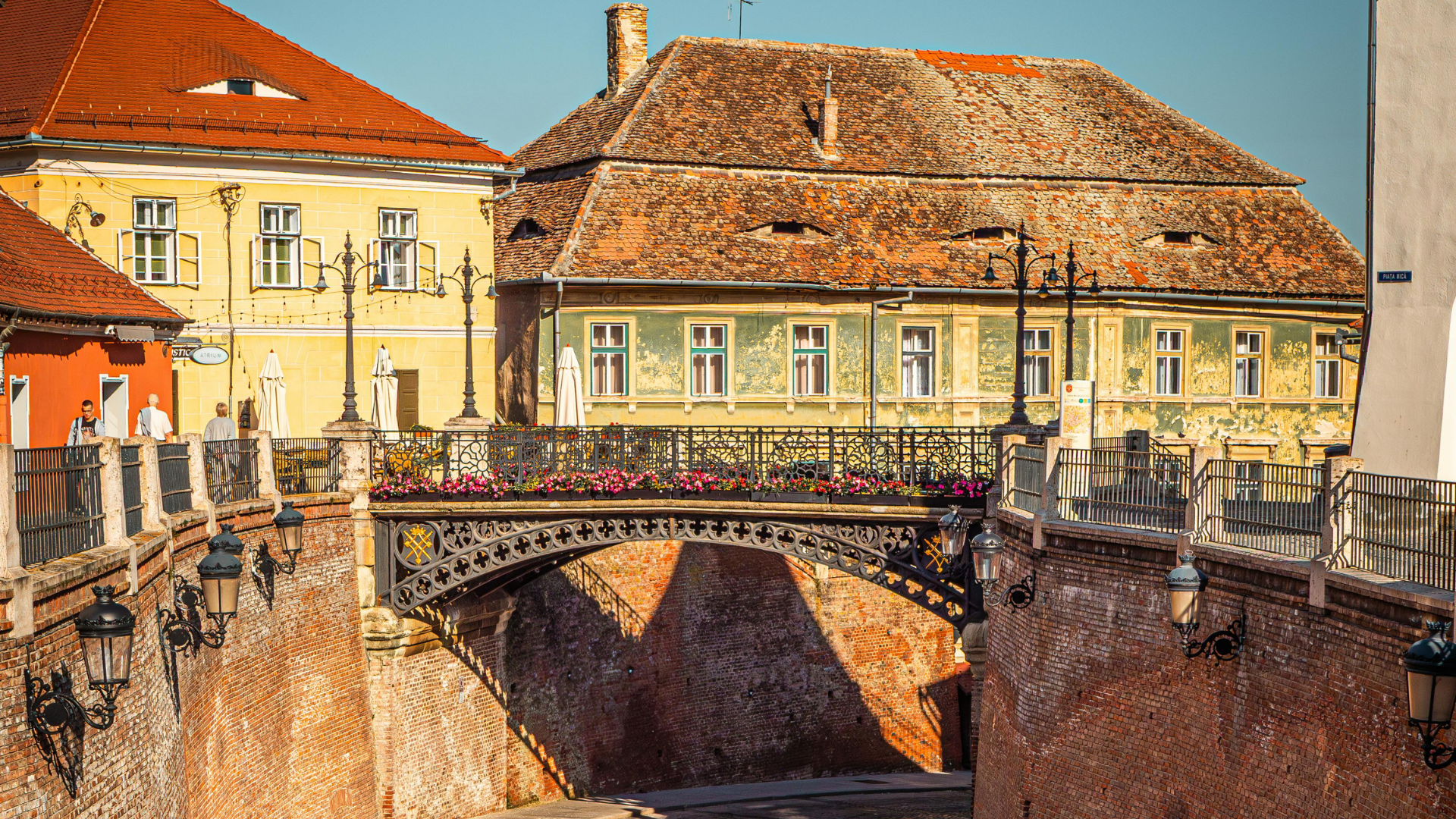 A city scene featuring old buildings with red tiled roofs, a decorative iron bridge with flower boxes, street lamps, and pedestrians walking along the sidewalk, with a clear blue sky overhead.
