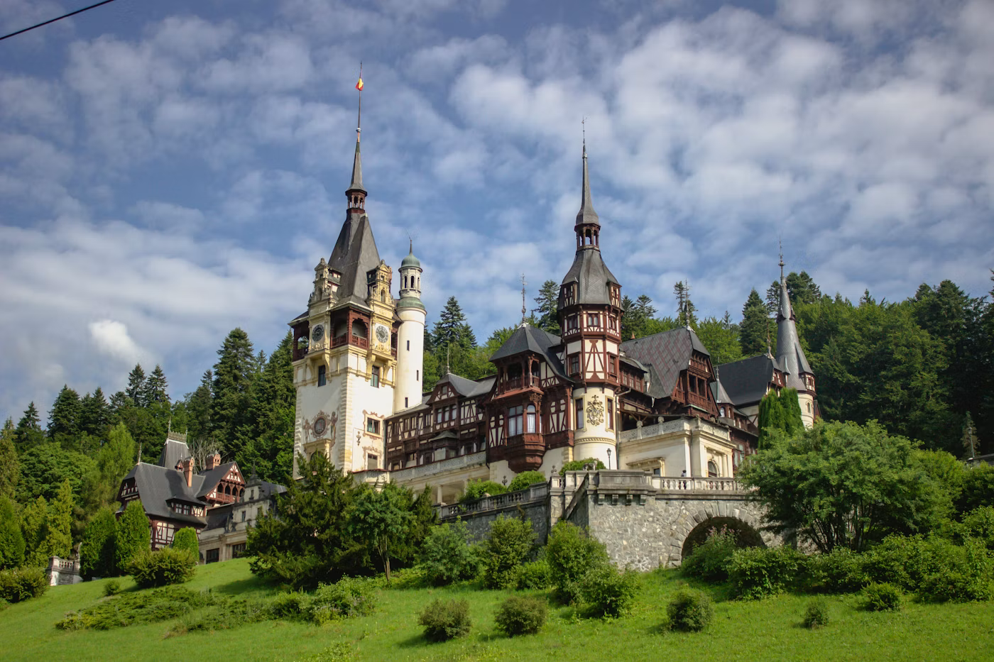 A large castle with multiple towers and spires, surrounded by green trees and grass, under a partly cloudy sky.
