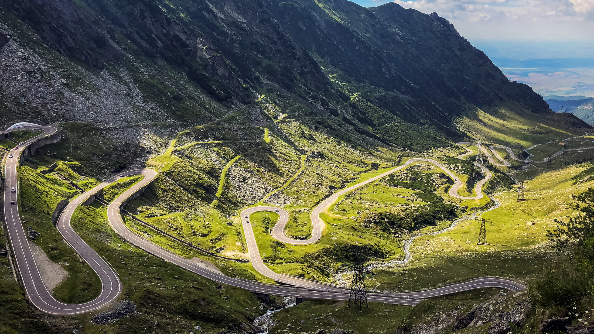 Winding mountain road with sharp turns running through green valley, with power lines and rocky mountain slopes in the background.