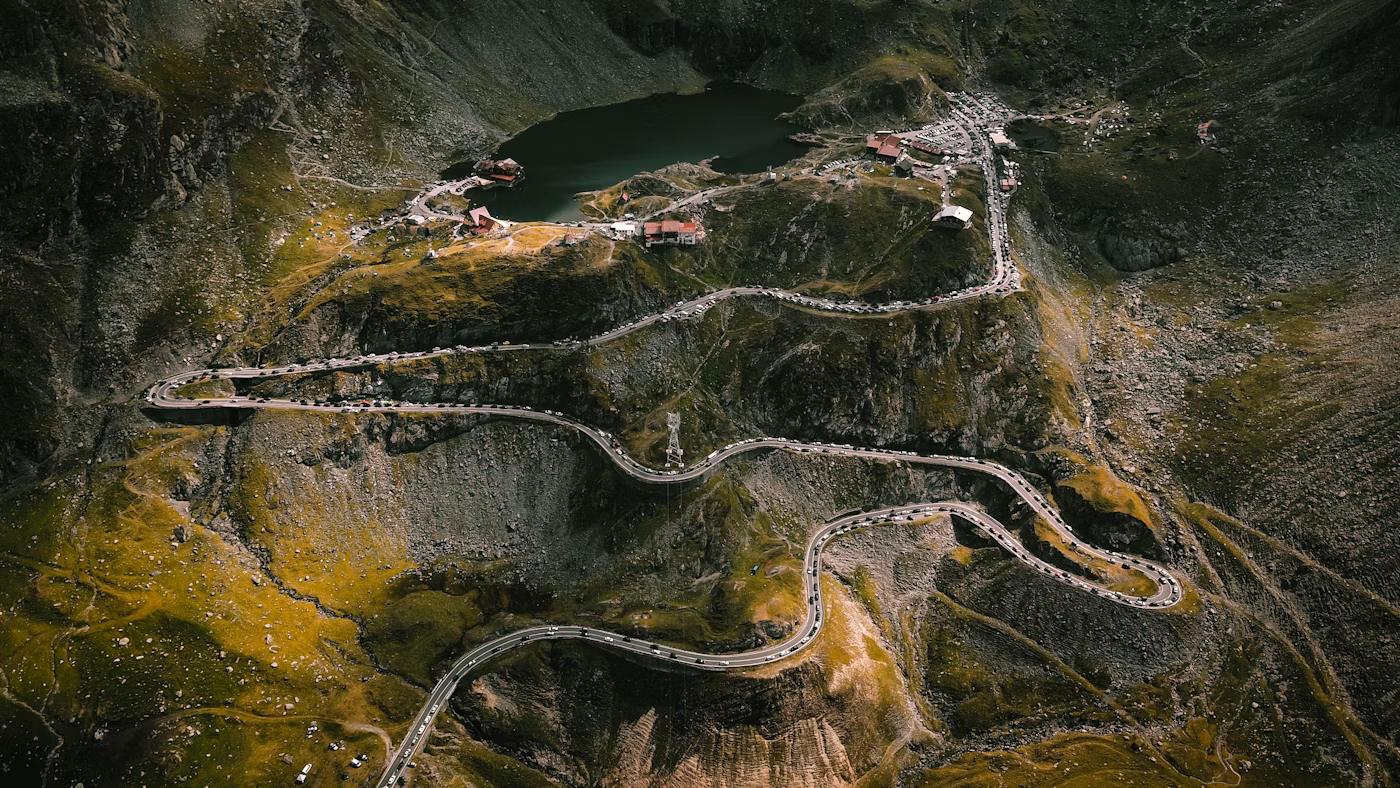 A winding mountain road runs through rugged terrain toward a small village with buildings and a lake at the top surrounded by steep cliffs.