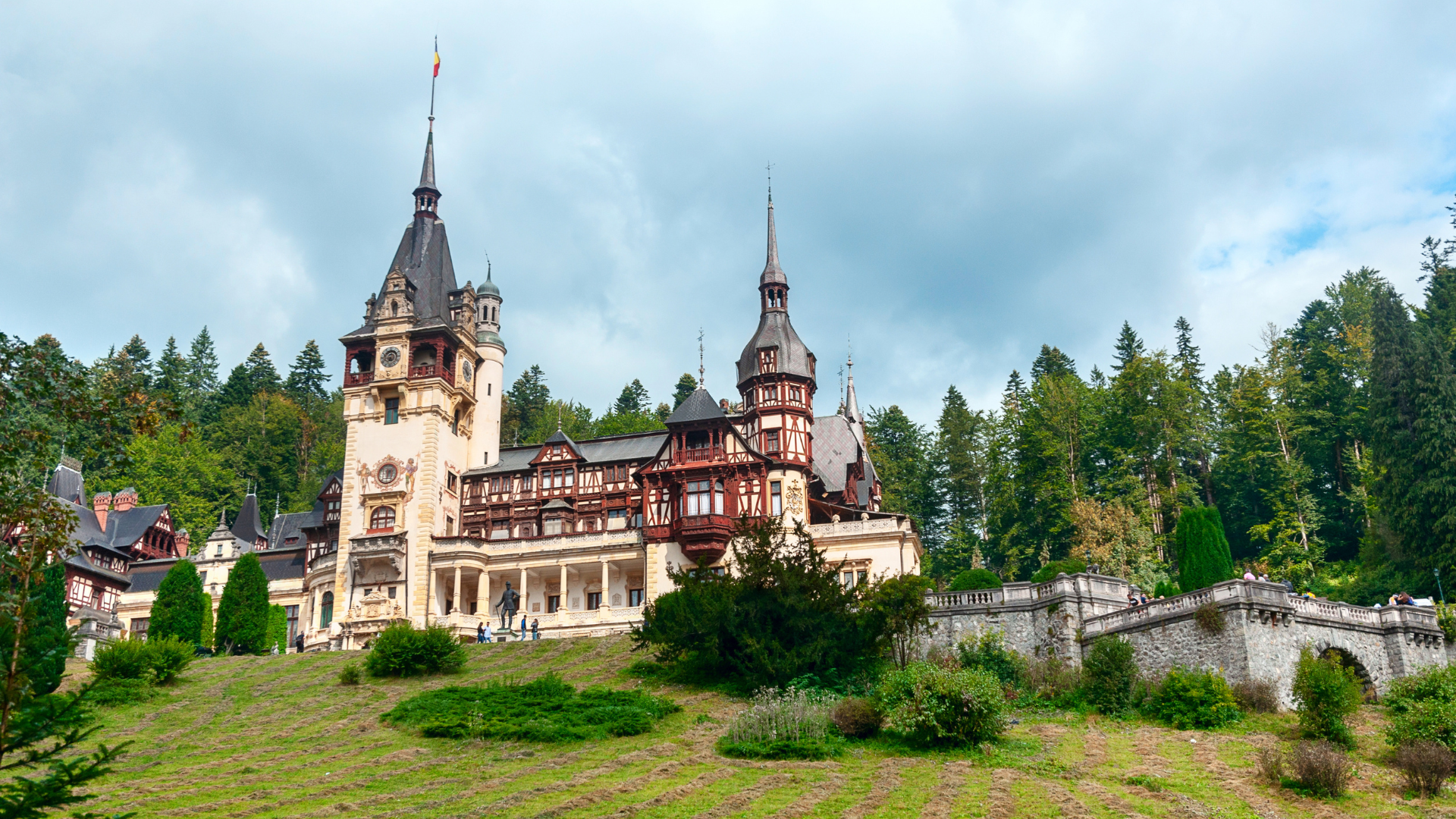 A large fairytale castle with tall spires, surrounded by trees and a garden with shrubs and plants, under a partly cloudy sky.