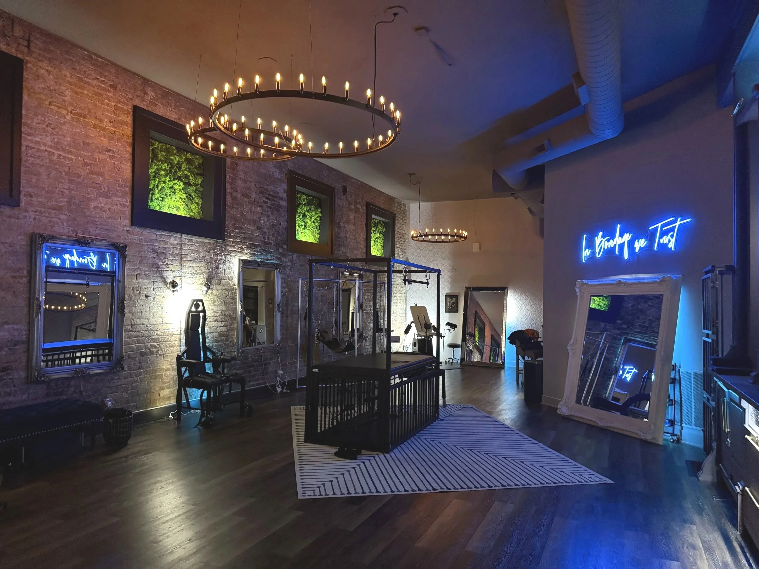 photography and bdsm studio with dim lighting, mirrored walls, wooden floor, modern bondage equipment, and a neon sign reading "In bondage we Trust." Chandeliers are overhead.