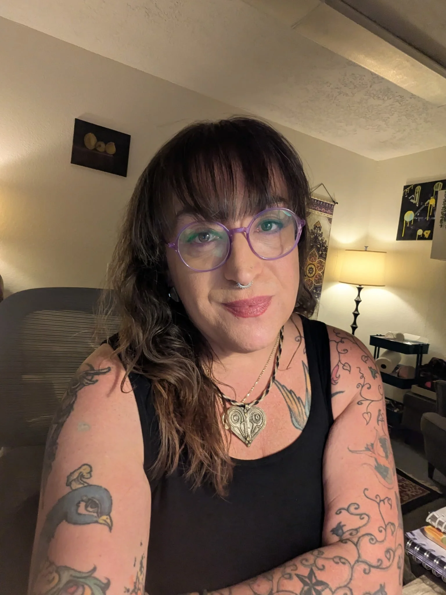 A woman with tattoos on her arms and chest, wearing purple glasses, a septum ring, and a black tank top, sitting in a warmly lit living room with artwork and a lamp in the background.
