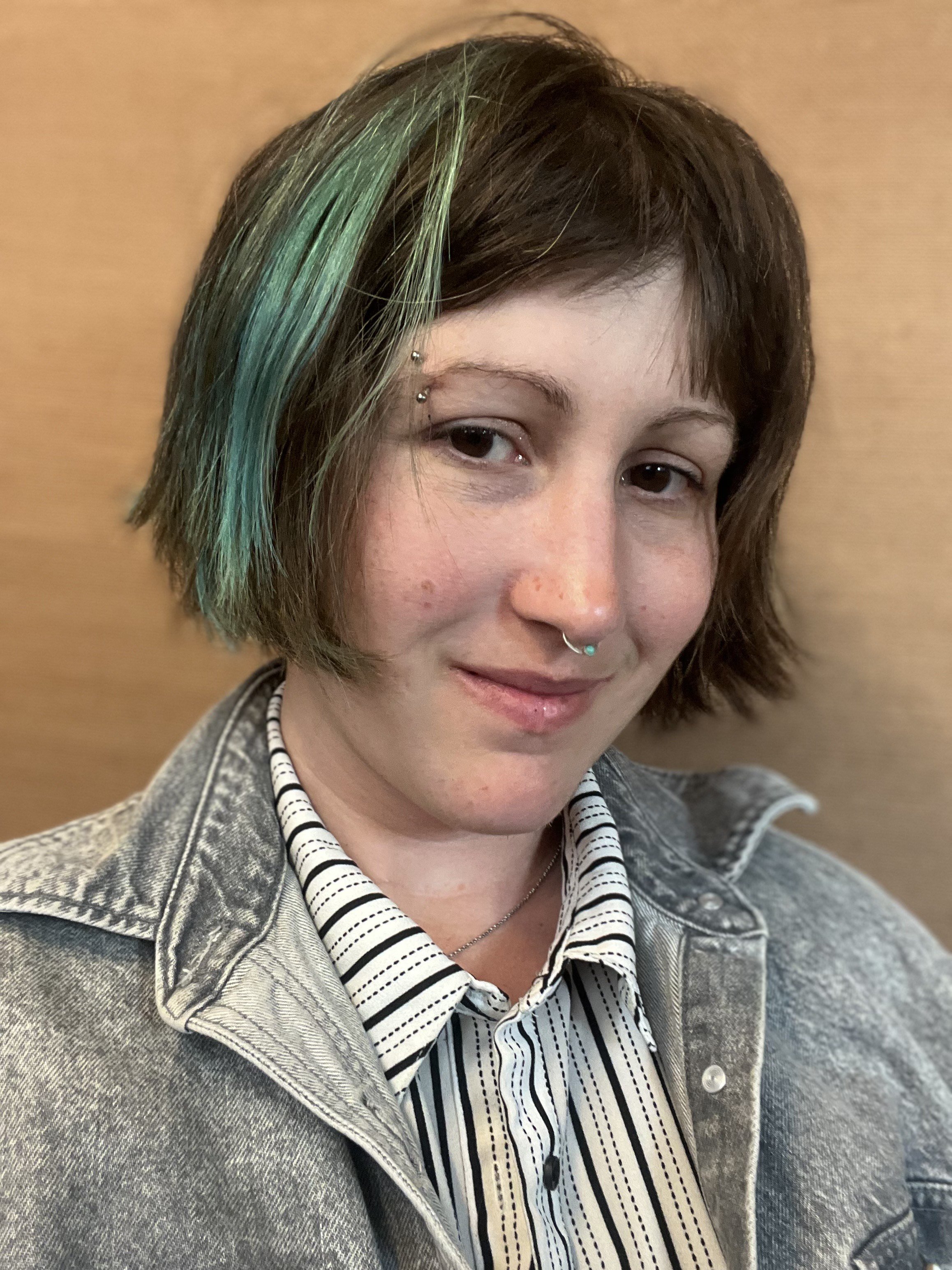 A young woman with short brown hair, some dyed turquoise, wearing a striped blouse and a denim jacket, standing against a wooden background.