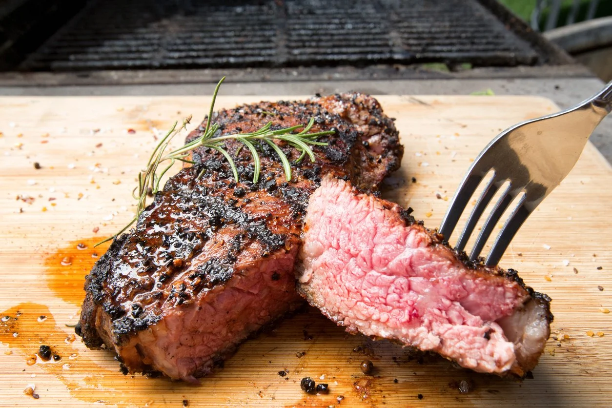Cooked sliced steak with a sprig of rosemary on a wooden cutting board, with grill in the background.