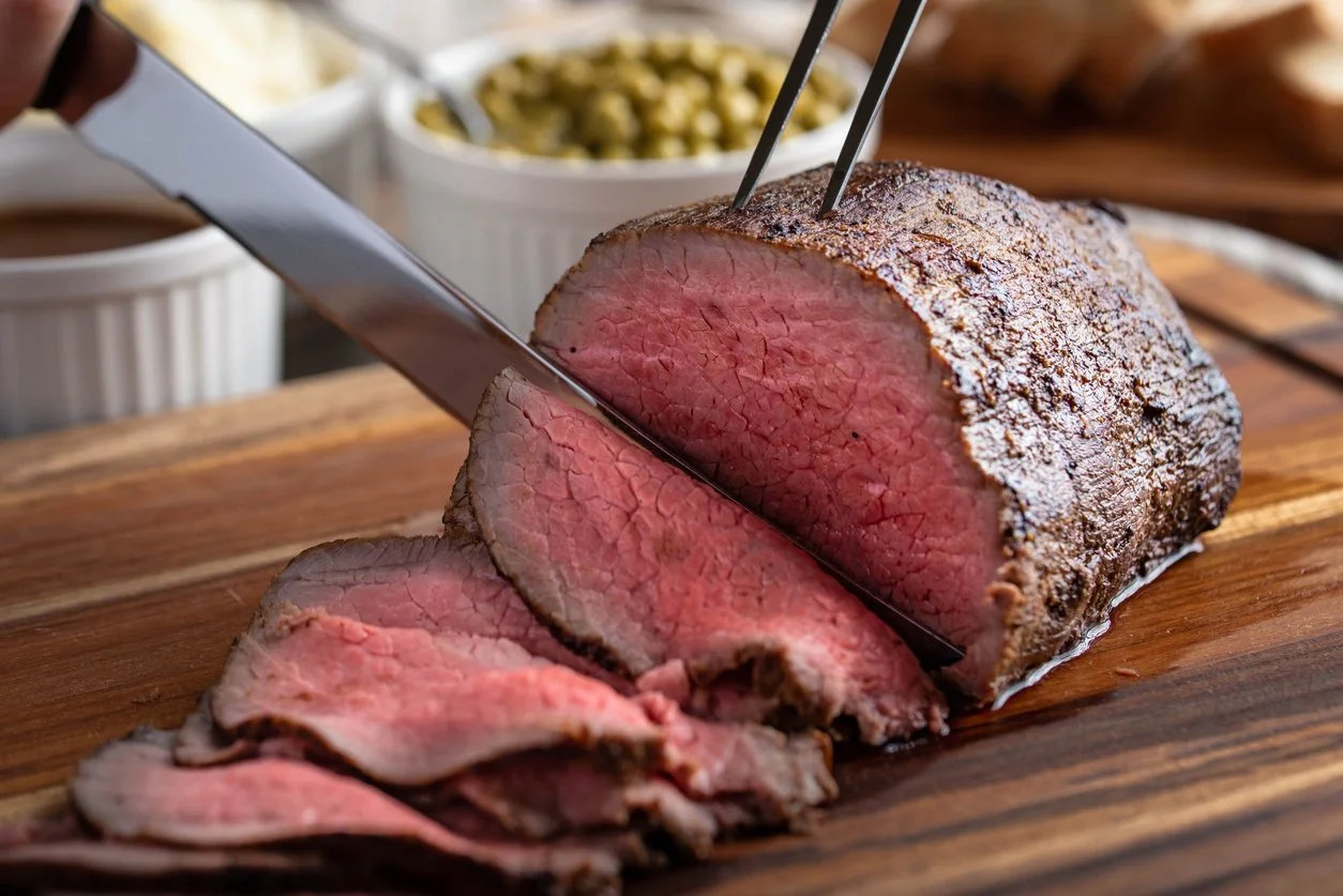 Sliced beef roast on a wooden cutting board with a sharp knife, with bowls of condiments in the background.