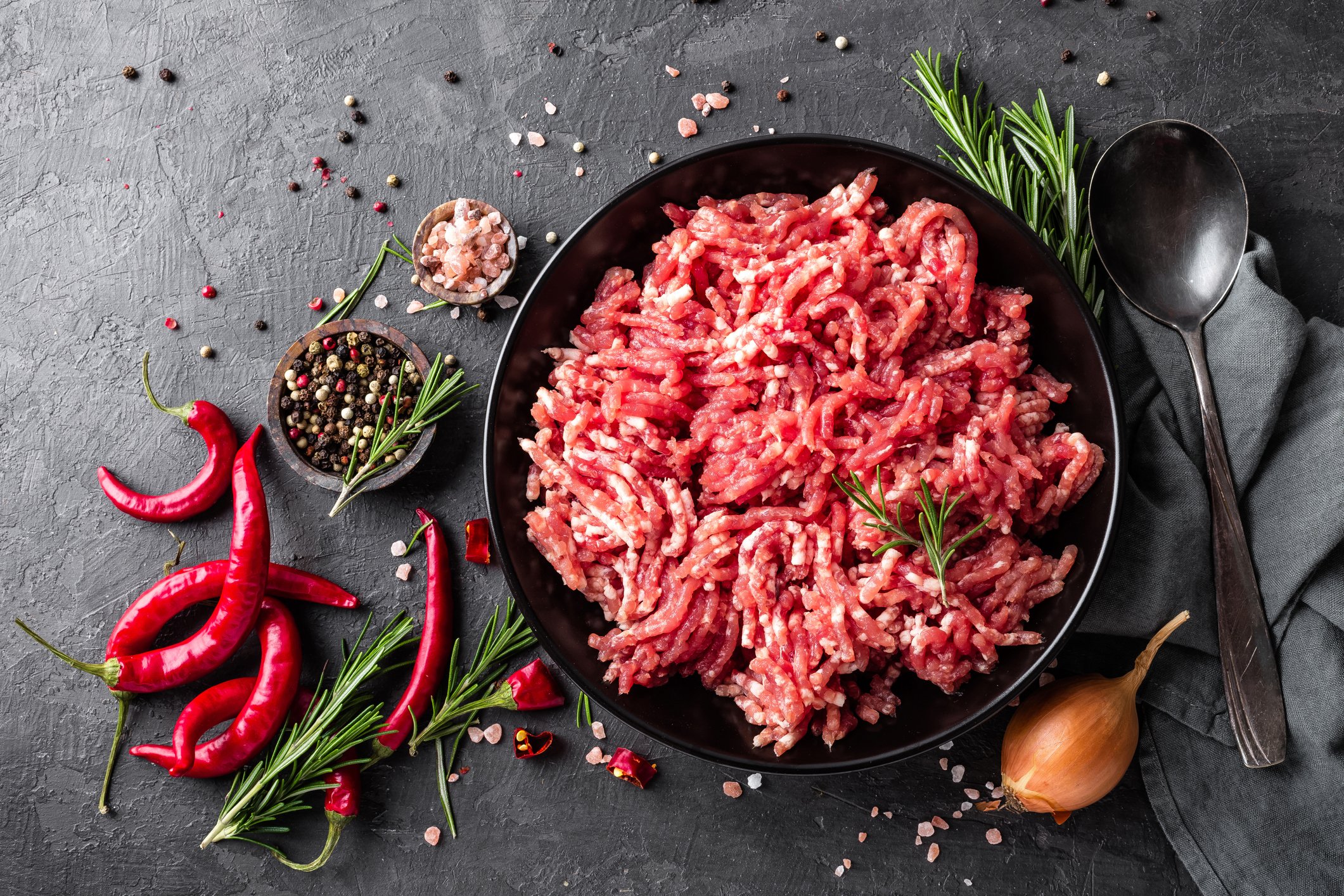 Raw ground meat in a black bowl, surrounded by red chili peppers, rosemary sprigs, salt, pepper, and a small garlic bulb on a dark textured surface.