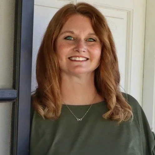 A woman with shoulder-length red hair, wearing a green top and a silver infinity necklace, smiling in front of a door.