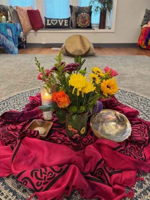 A bouquet of colorful flowers in a vase on a red cloth, surrounded by a candle and a shell, with a cozy living room in the background featuring pillows and a