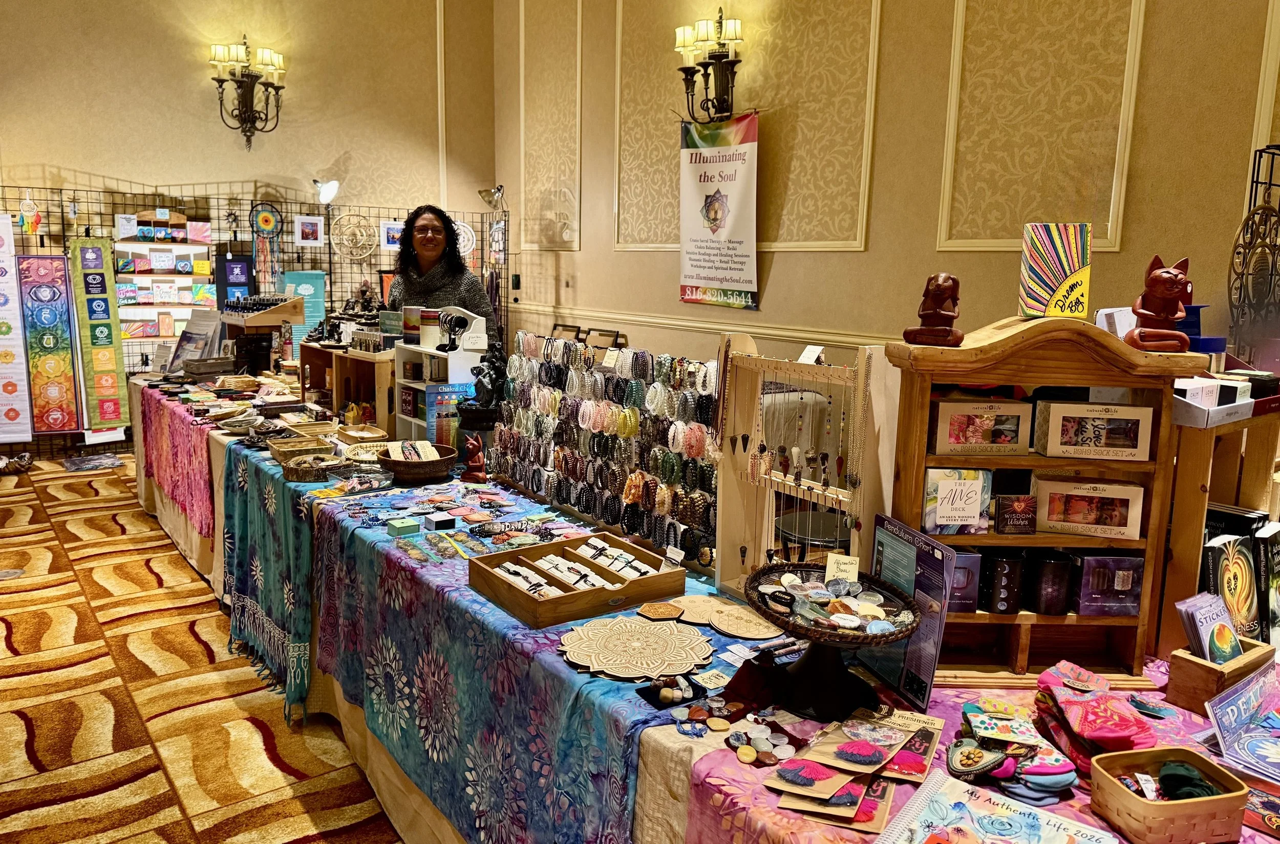 A woman with glasses behind a vendor table selling jewelry and spiritual items at an indoor market. The table is covered with a colorful cloth and displays various bracelets, cards, and small items. Behind her, there are more tables with similar merc