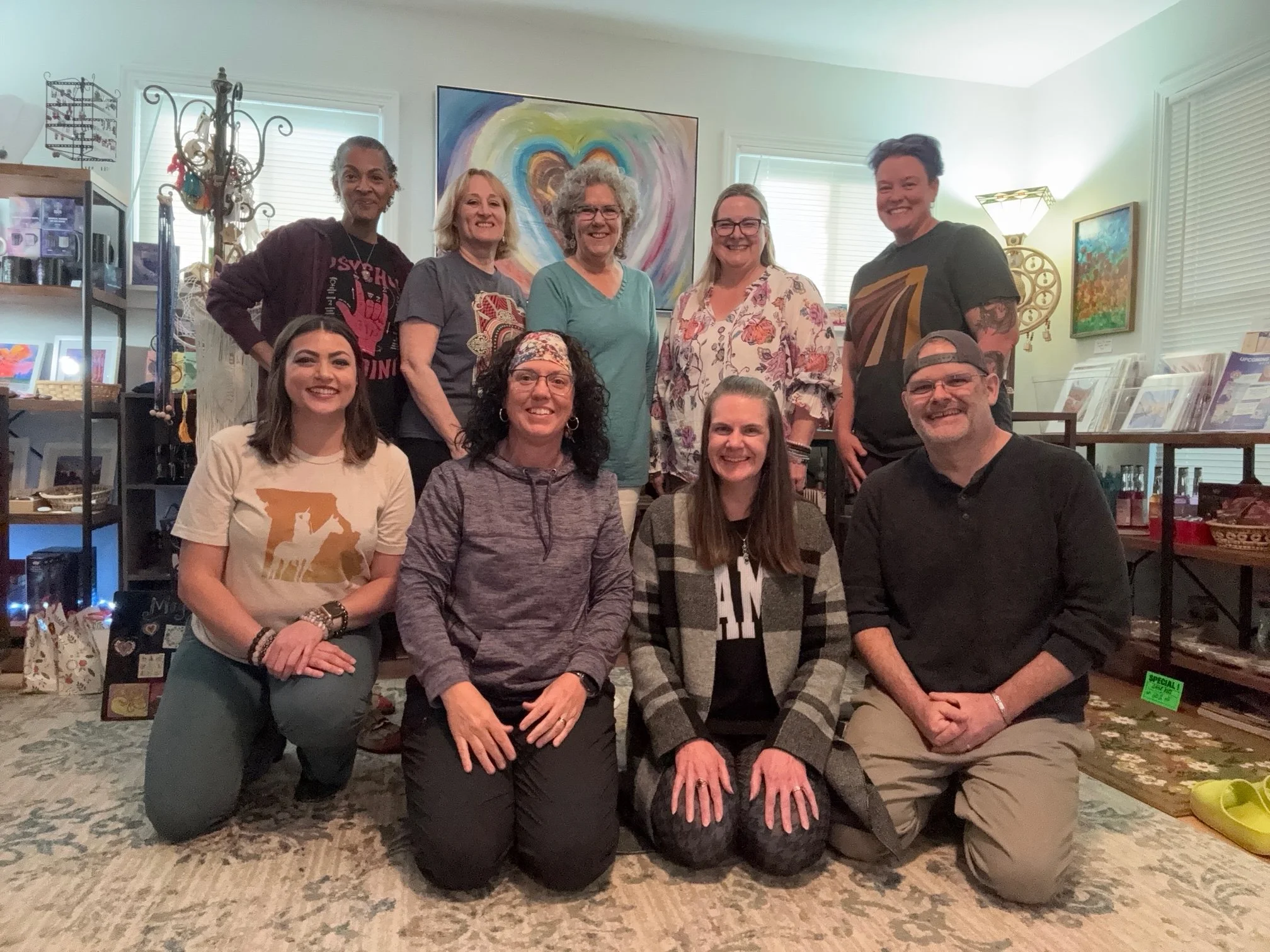 A group of ten women and one man posing in an art gallery or shop. They are smiling, with some kneeling and some standing. Behind them, modern colorful artwork and shelves with various framed pictures and items are visible. The room has white walls a