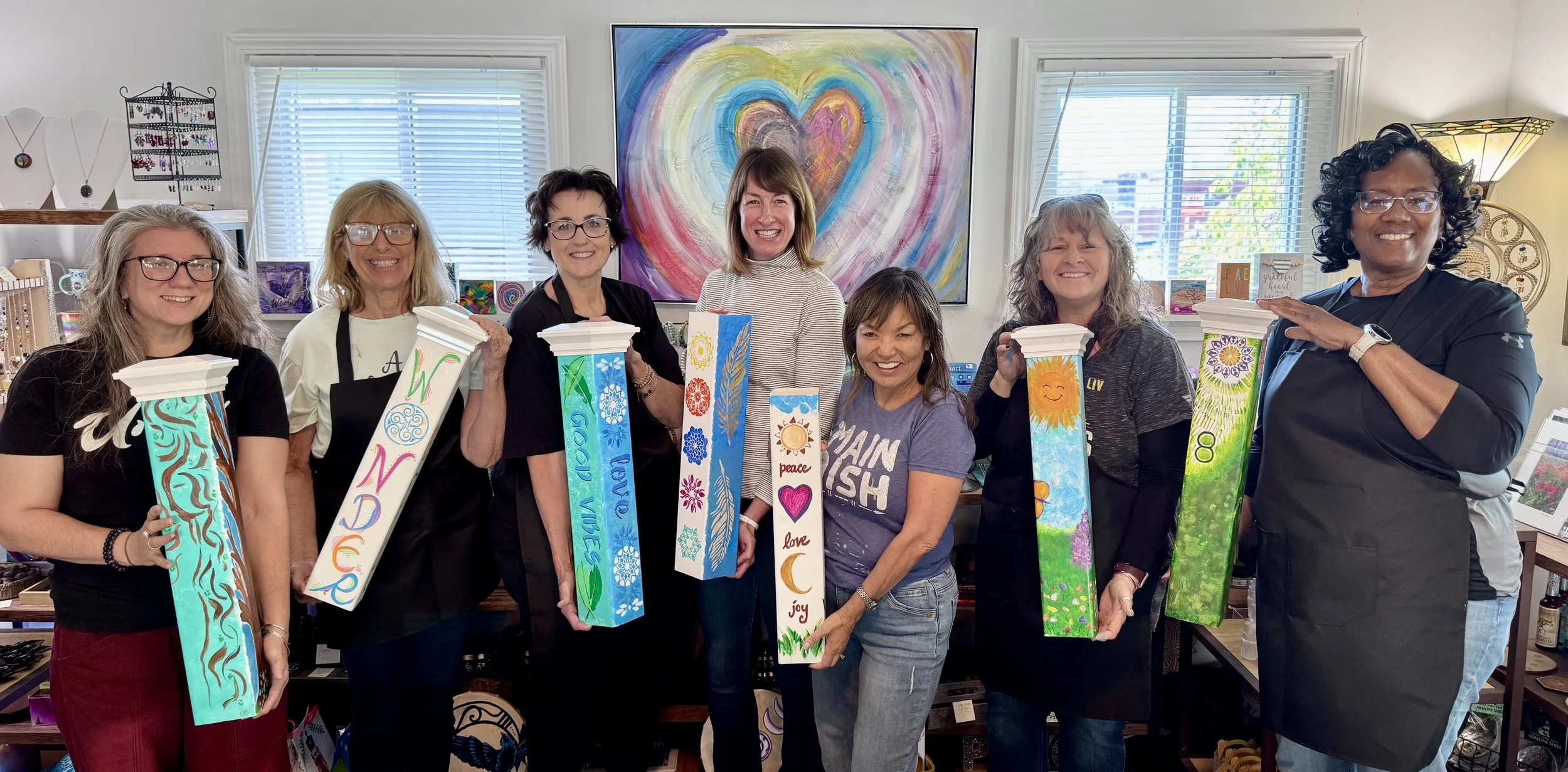 Eight women standing in an art studio, holding painted wooden blocks with colorful designs and words like 'wonder,' 'peace,' and 'joy.' They are smiling and appear happy, with a large colorful heart painting on the wall behind them.