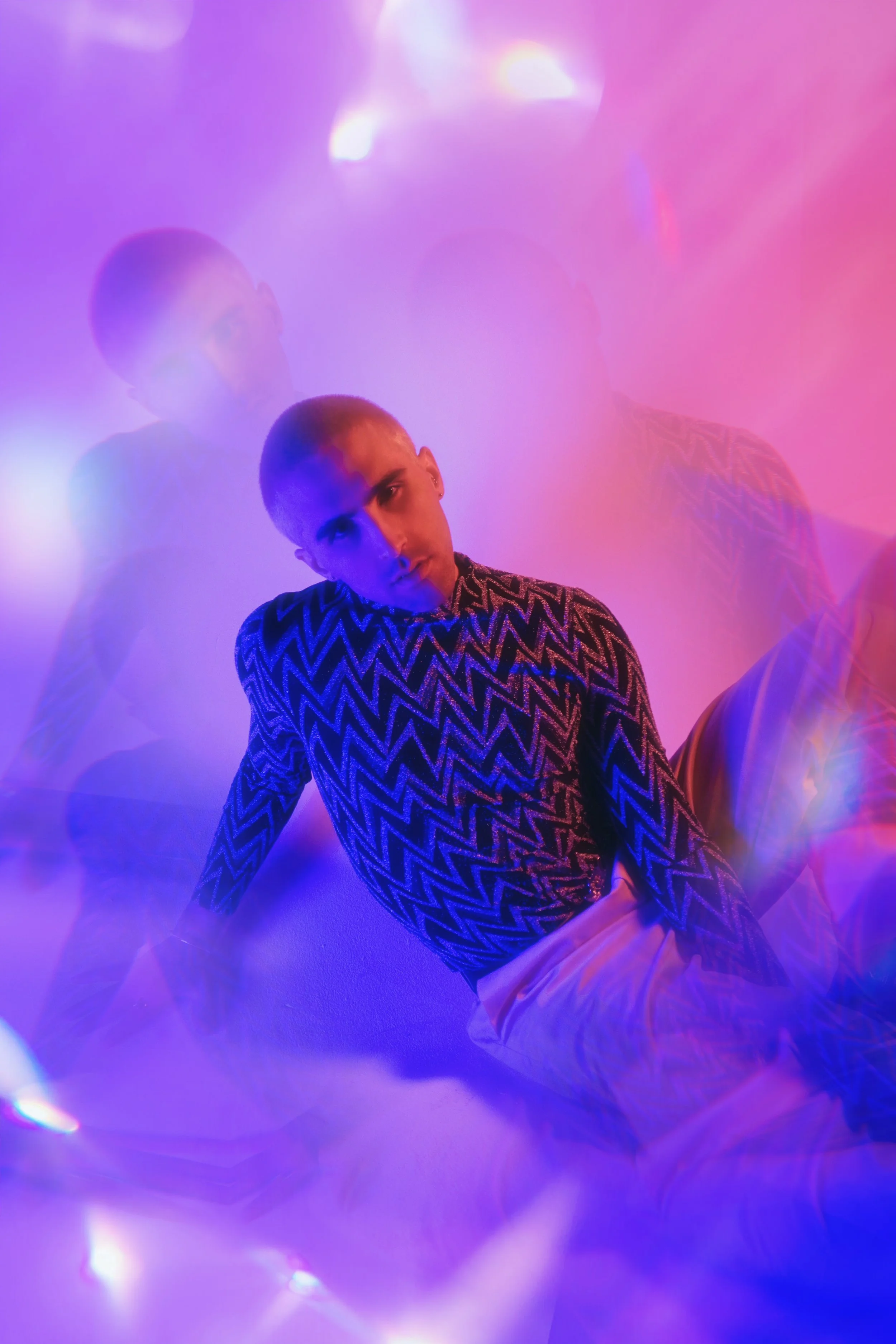 A person with a close-cropped hairstyle wearing a patterned shirt, seated on the floor, surrounded by colorful pink, purple, and blue lights creating a dreamy, hazy atmosphere.