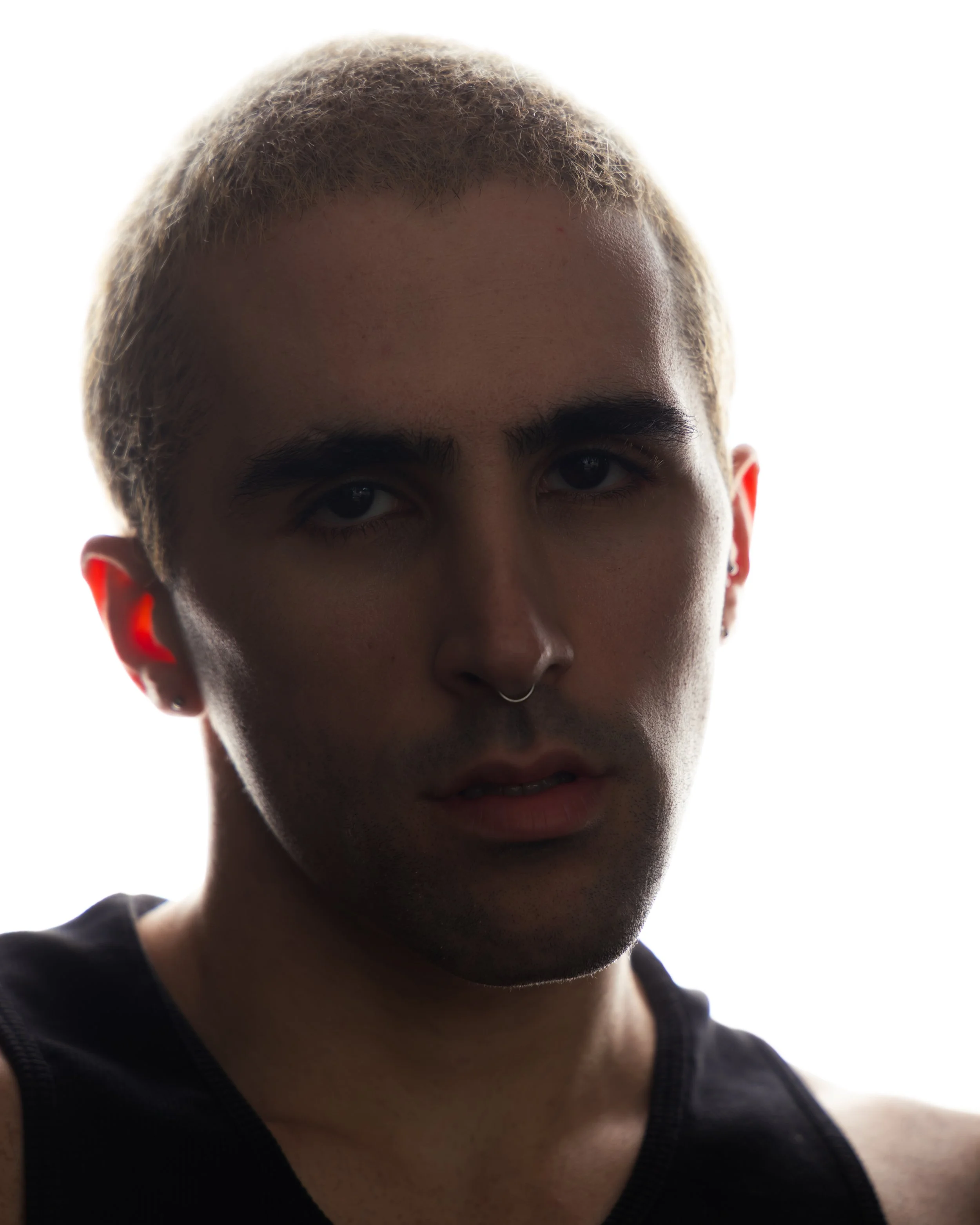 Portrait of a young man with short, light-colored hair and dark eyebrows, wearing a studded septum ring and a sleeveless black shirt, against a bright, white background.