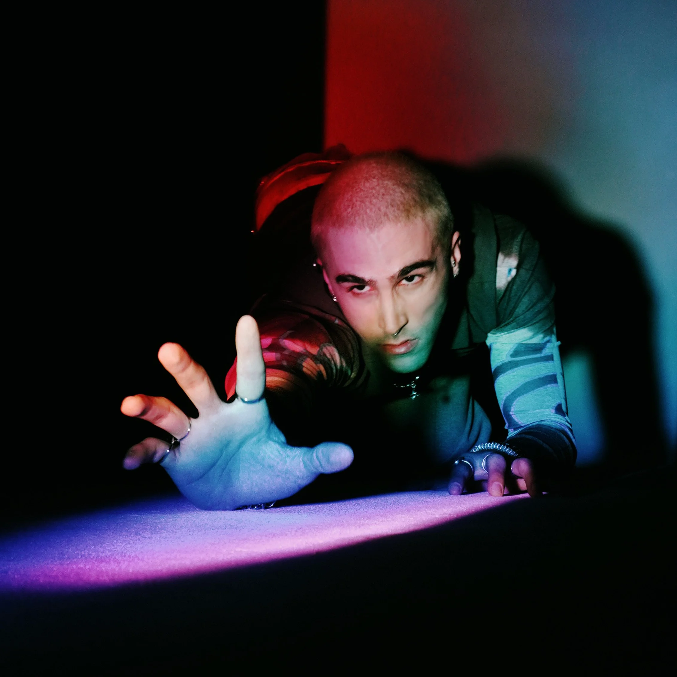 A person with short hair reaching towards the camera, wearing rings, earrings, and layered clothing, with multicolored lighting creating a dramatic effect.