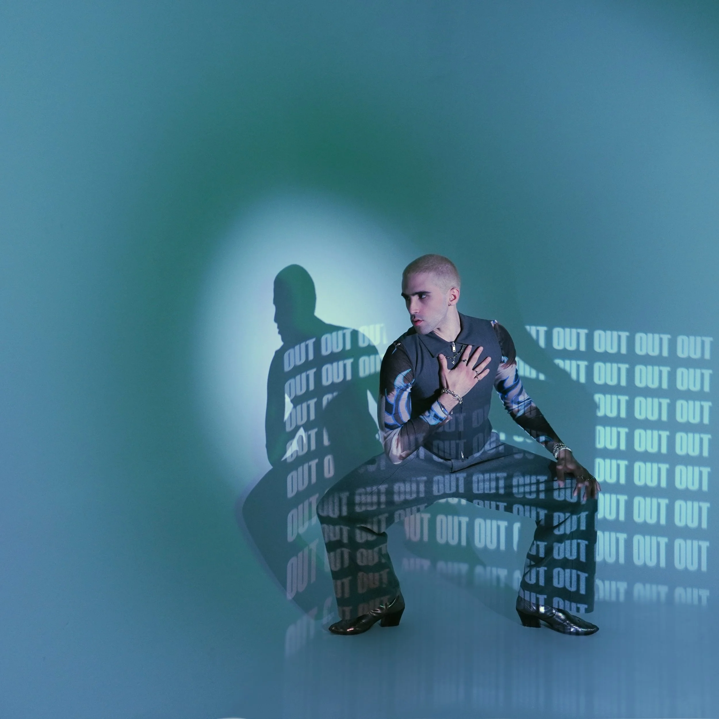 A person in fashionable clothing sitting on the floor with one hand on their chest and the other on their knee, with a shadow cast on the wall behind them. The shadow has the repeated word "OUT" projected onto it.