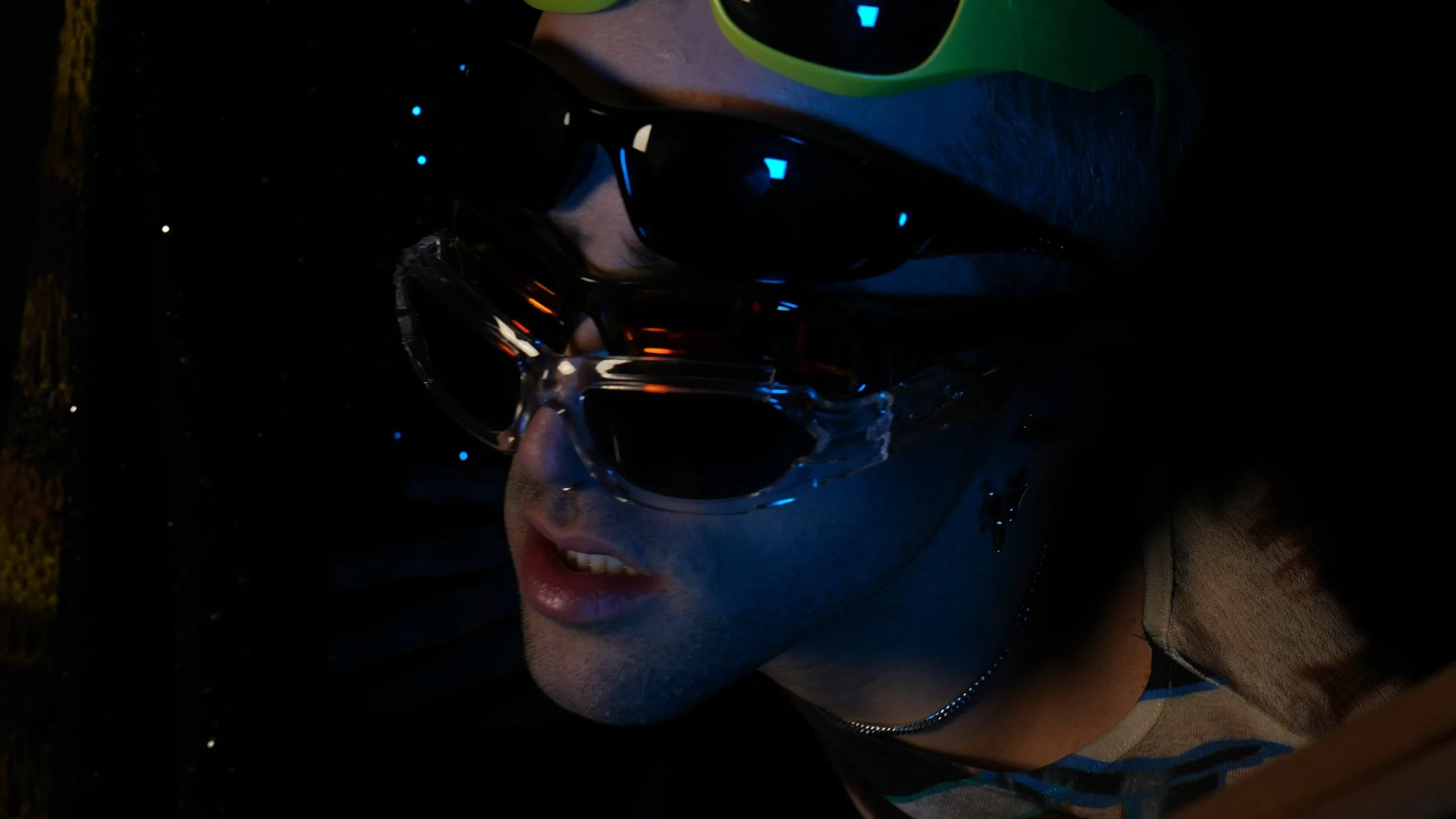 Close-up of a young man wearing multiple pairs of sunglasses, with dim lighting and a dark background.