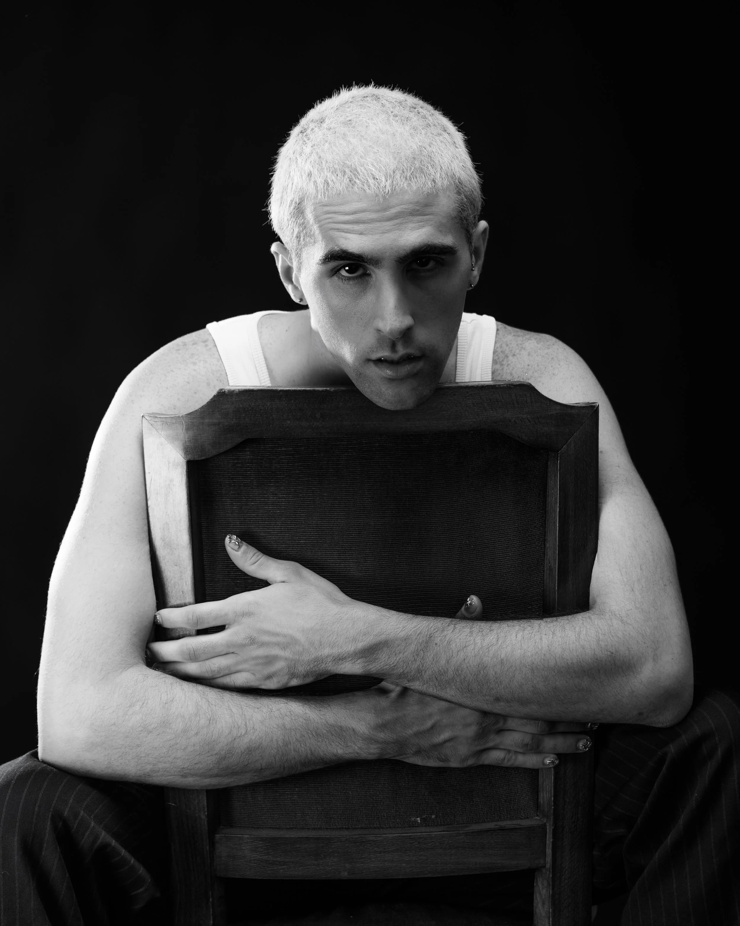 Black and white portrait of a person with platinum blonde hair, sitting on a chair and hugging a wooden framed chair back, with intense expression, in a studio setting.