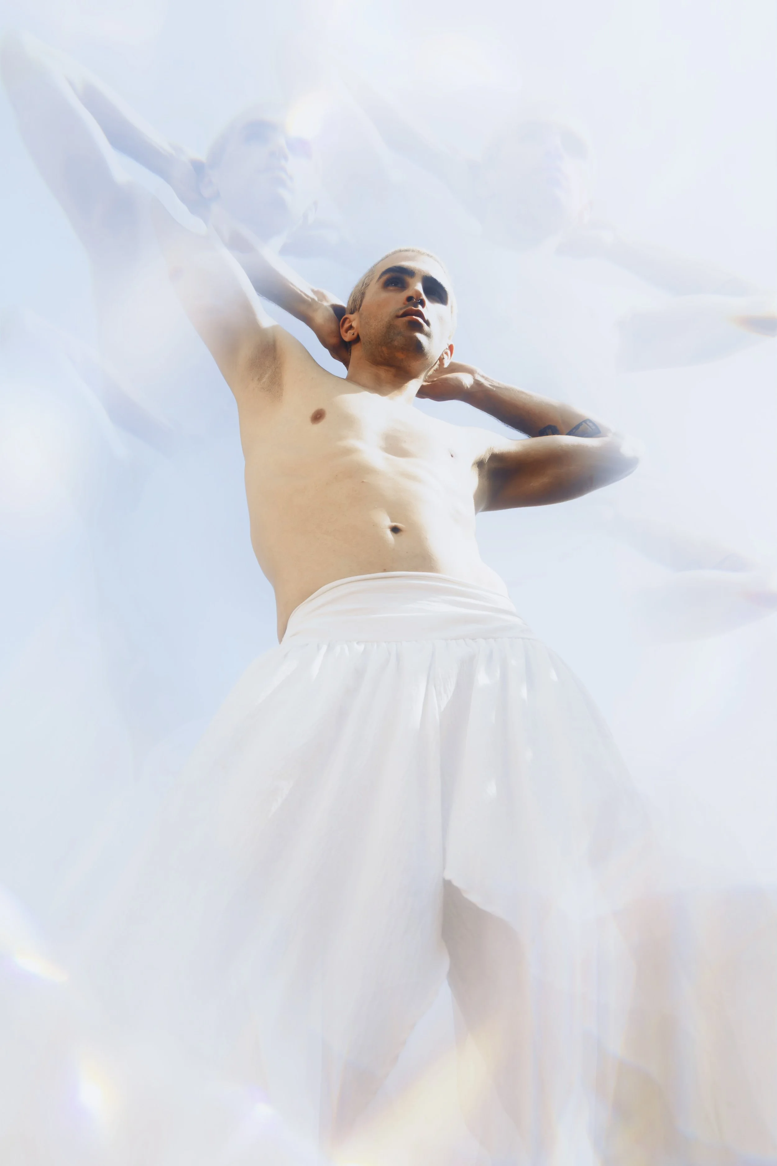 A shirtless man with short hair and wearing white pants, standing with arms behind his head against a bright sky background, with a semi-transparent duplicate of himself overlapping in a dreamy effect.